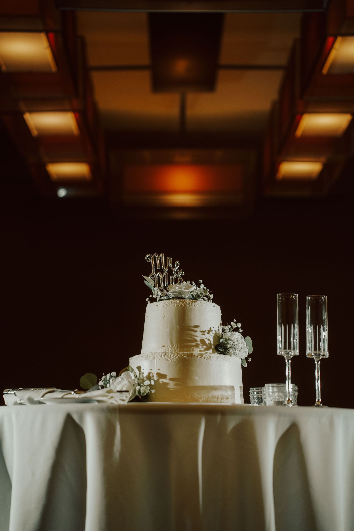 A white wedding cake sits elegantly on a table with soft lighting and glassware in a dark reception venue.
