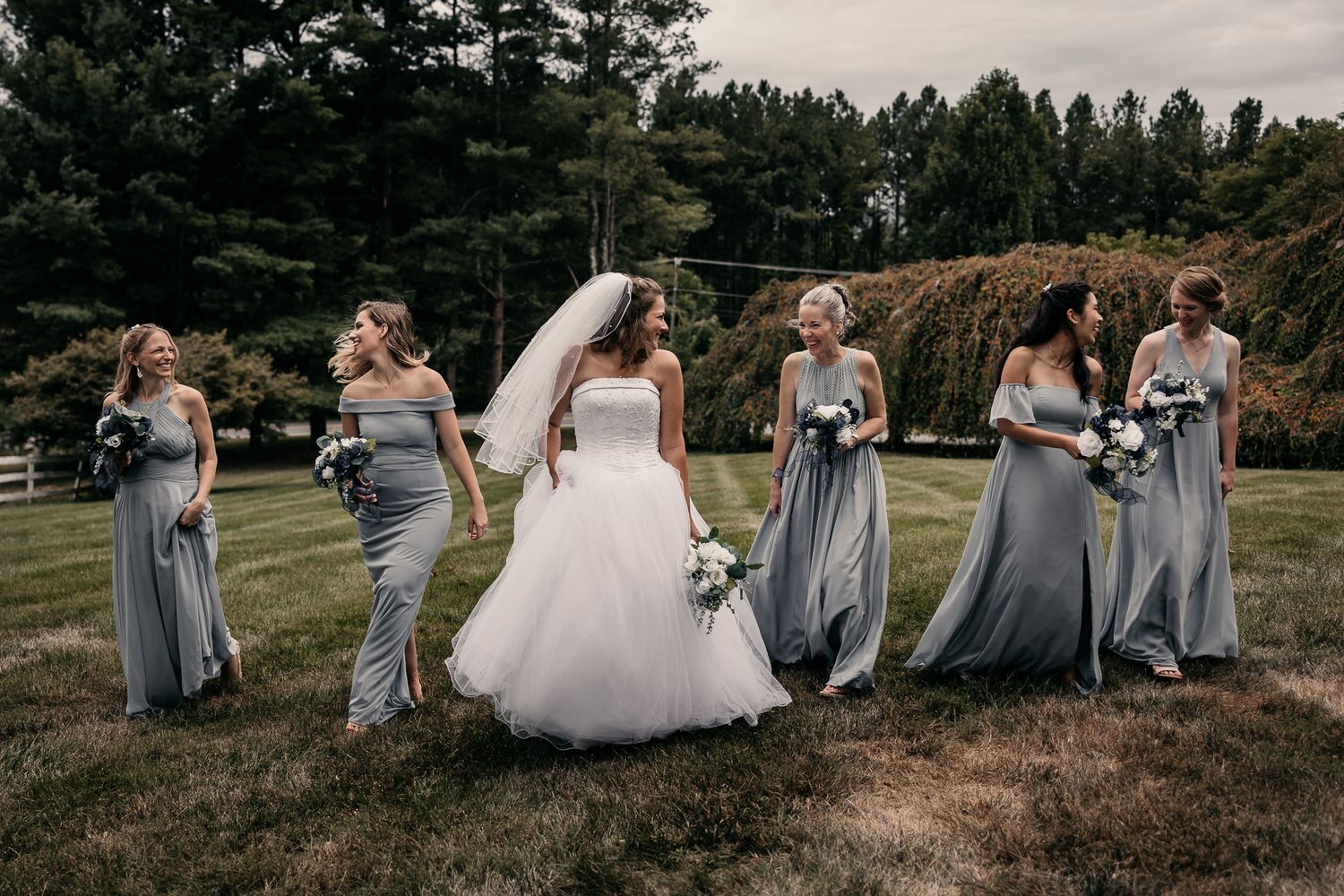 A bridal party in gray dresses and a white wedding gown pose together on a grassy hill with evergreen trees in the background.