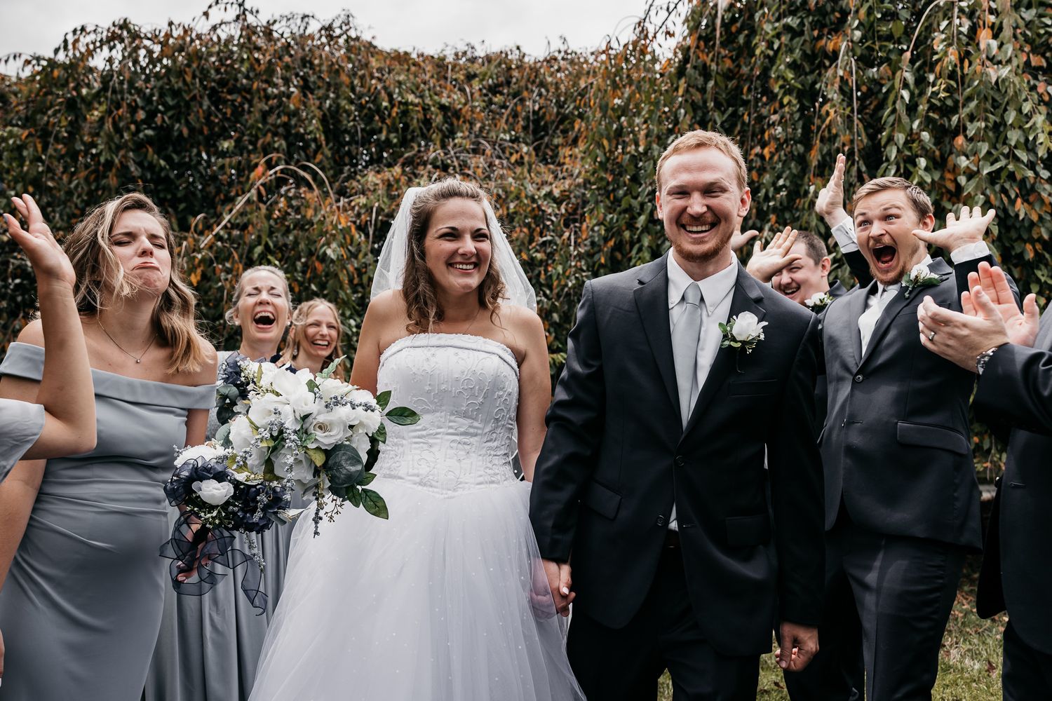 Wedding party in gray and black attire laughs together outdoors with willow trees in background.