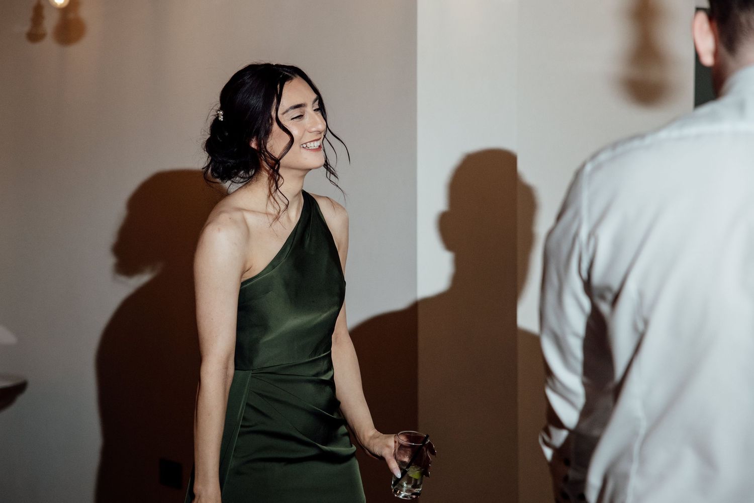Elegant woman in emerald green satin dress laughs joyfully while holding drink at wedding reception.