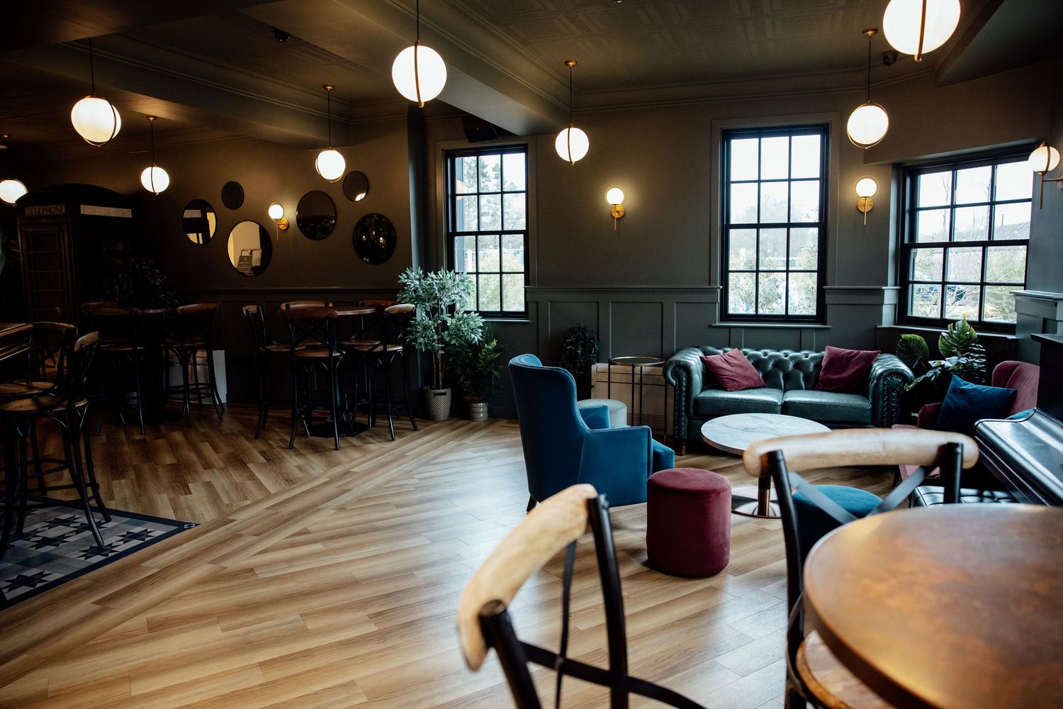 Elegant bar lounge with hardwood floors, pendant lights, velvet sofas, and high bar seating in a moody, stylish interior.
