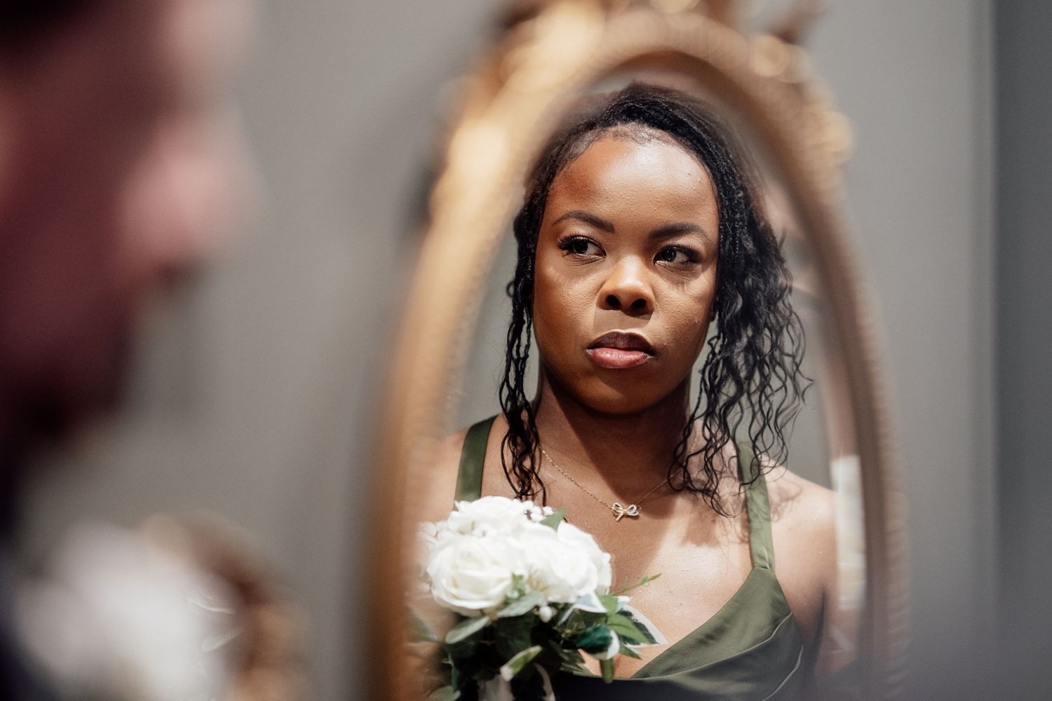 Elegant bridesmaid in olive dress holding white roses reflected in ornate mirror at wedding.