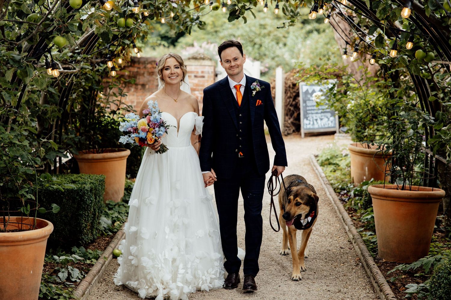Garden pathway lined with potted plants and string lights for a wedding ceremony.