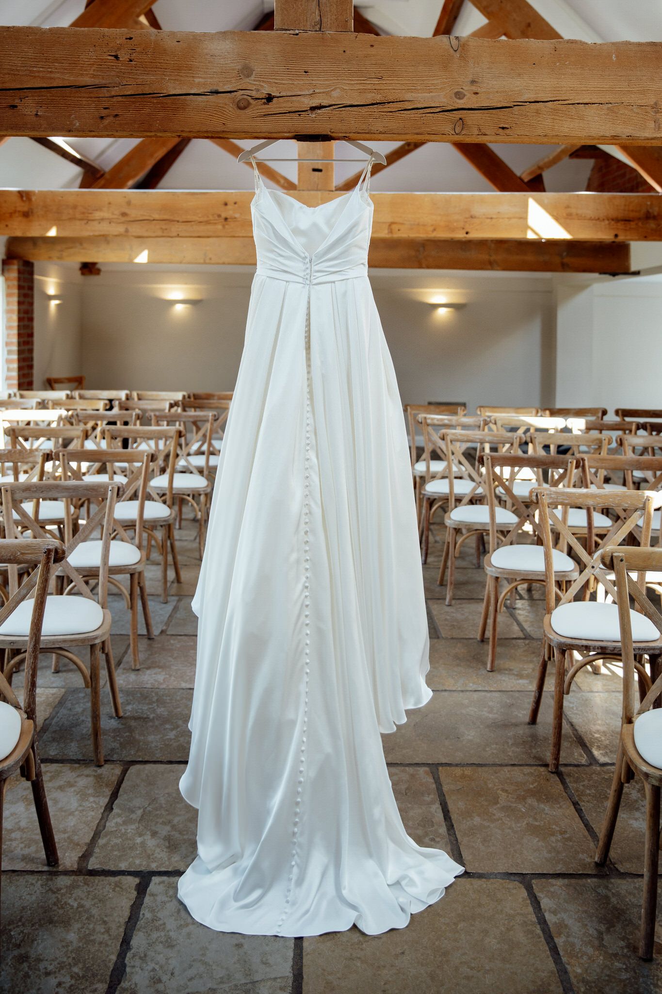 White wedding dress displayed in a rustic barn venue with wooden beams and cross-back chairs.