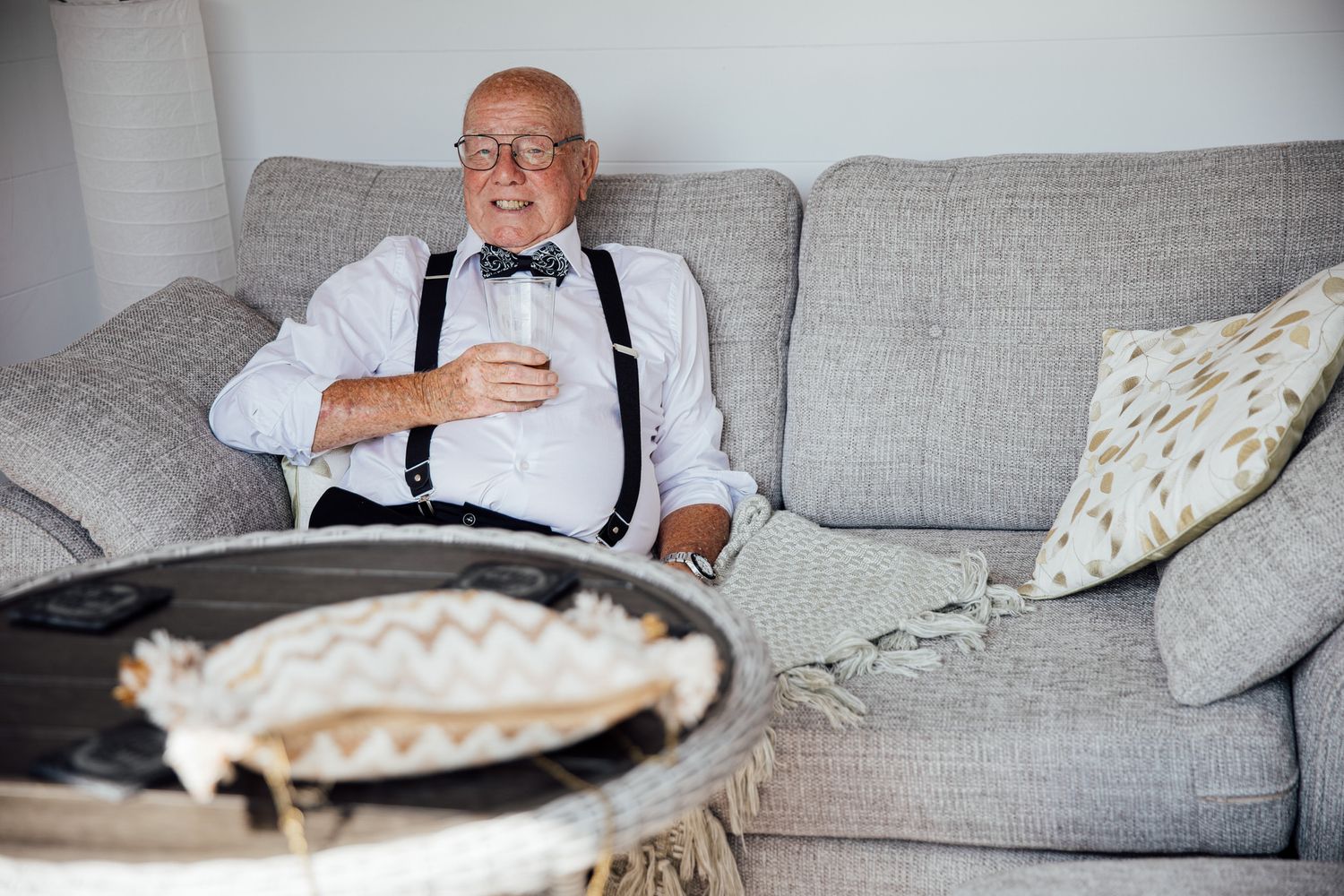 Elderly man in suspenders and bowtie relaxing on grey couch during wedding preparations.
