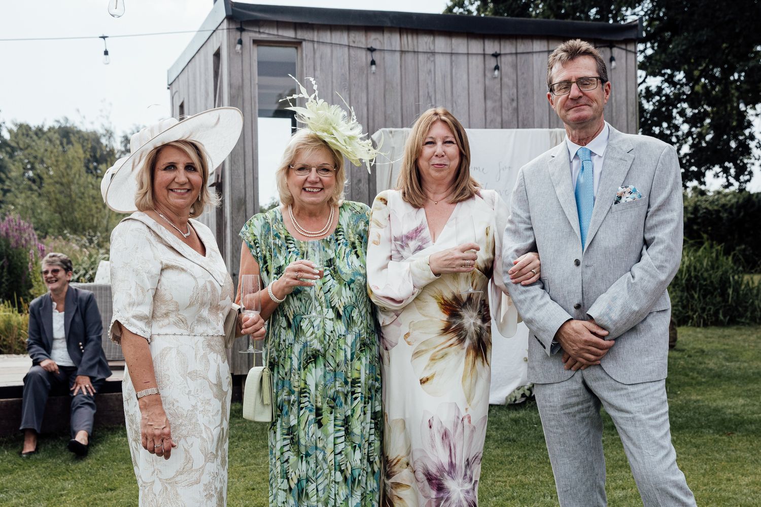 Wedding guests in formal attire pose together outdoors near a rustic building.