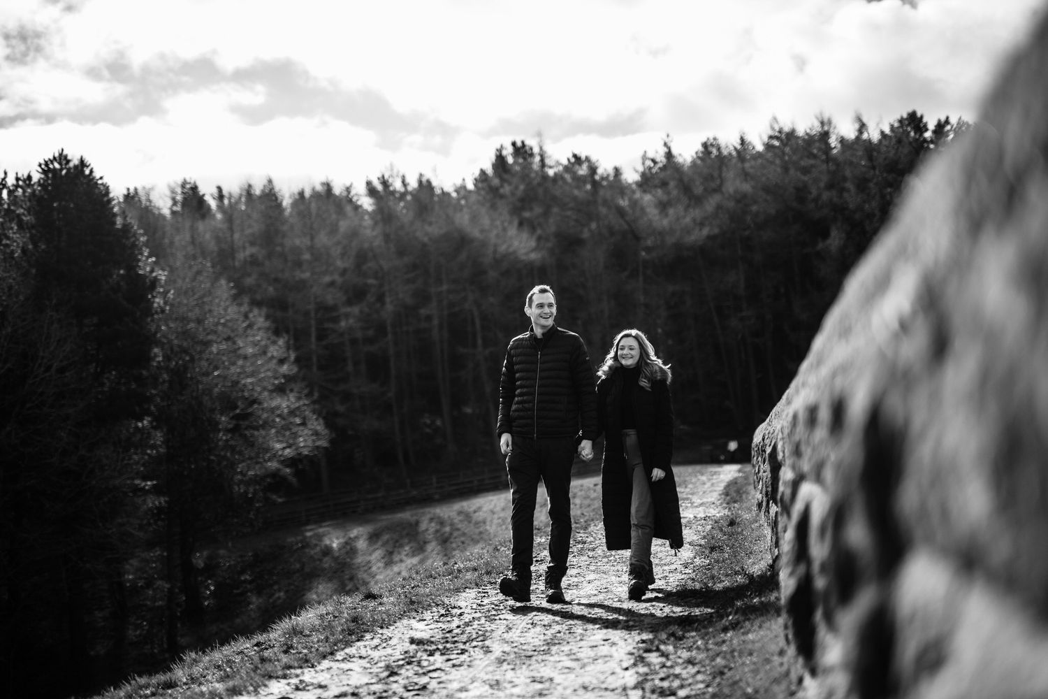 Two people walk hand in hand on a forest path with evergreen trees in the background.