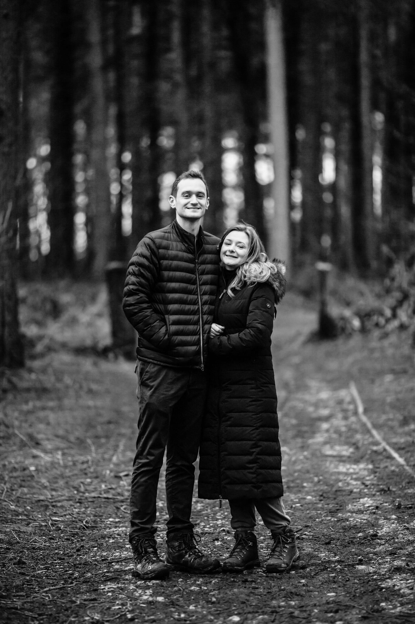 Black and white portrait of couple in winter coats standing together in wooded setting.