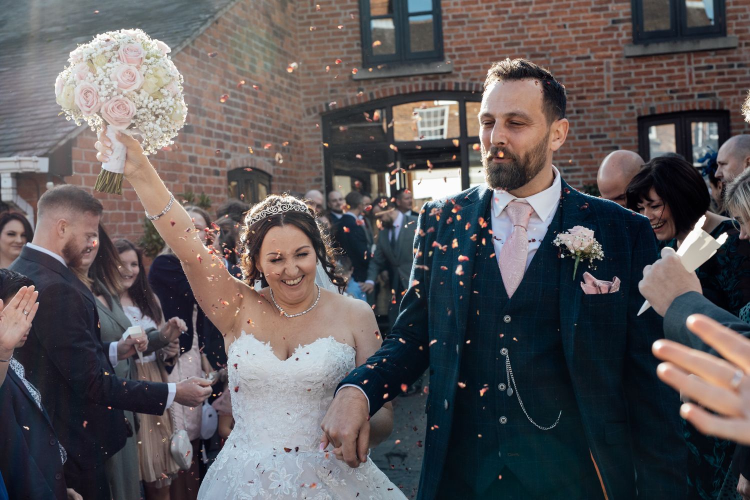 Wedding party celebrates with confetti toss outside brick venue during daytime festivities.