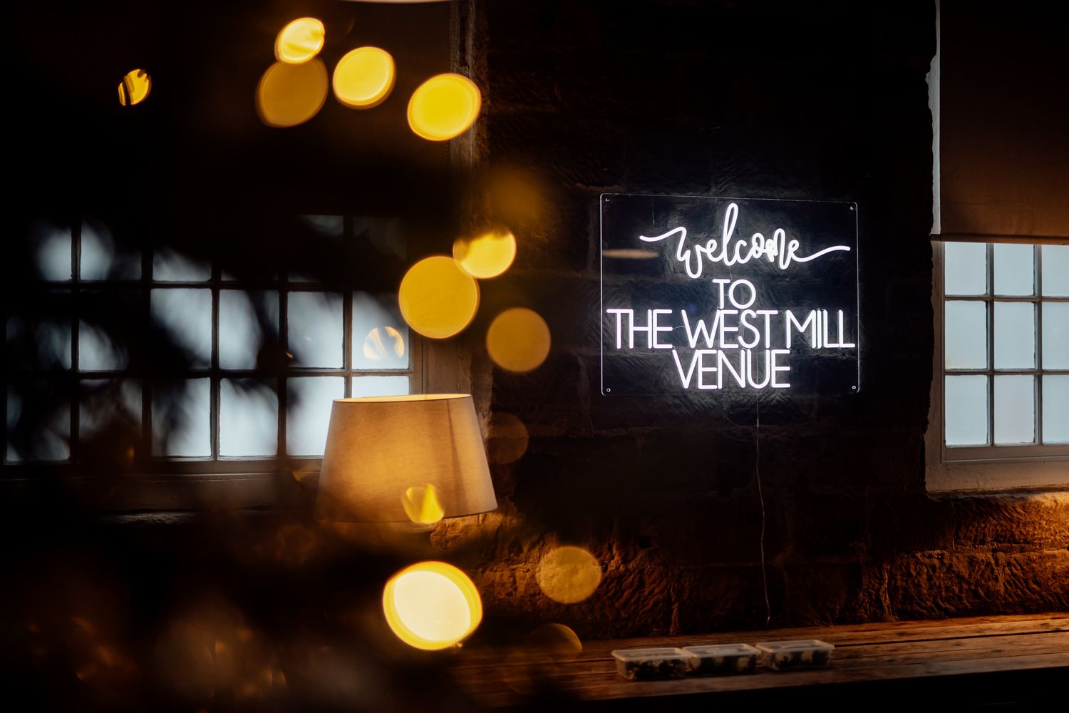 Warm bokeh lights create an atmospheric glow against a dark background at The West Mill venue.