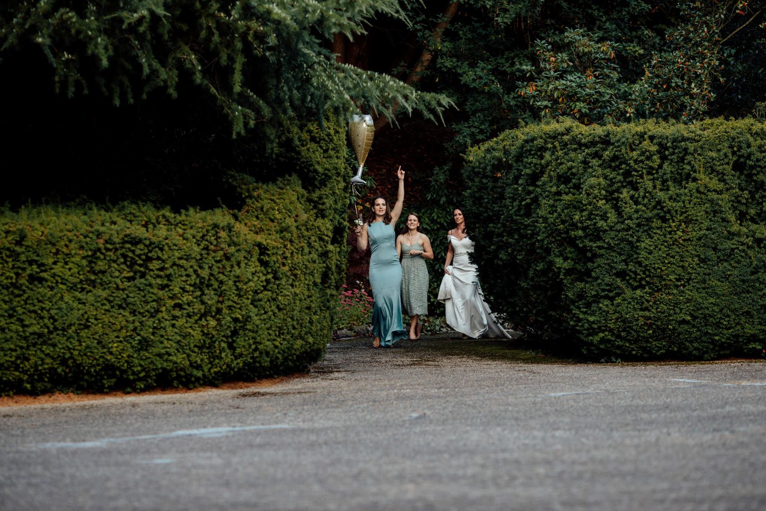 Wedding party walking along a tree-lined path in a lush garden setting.