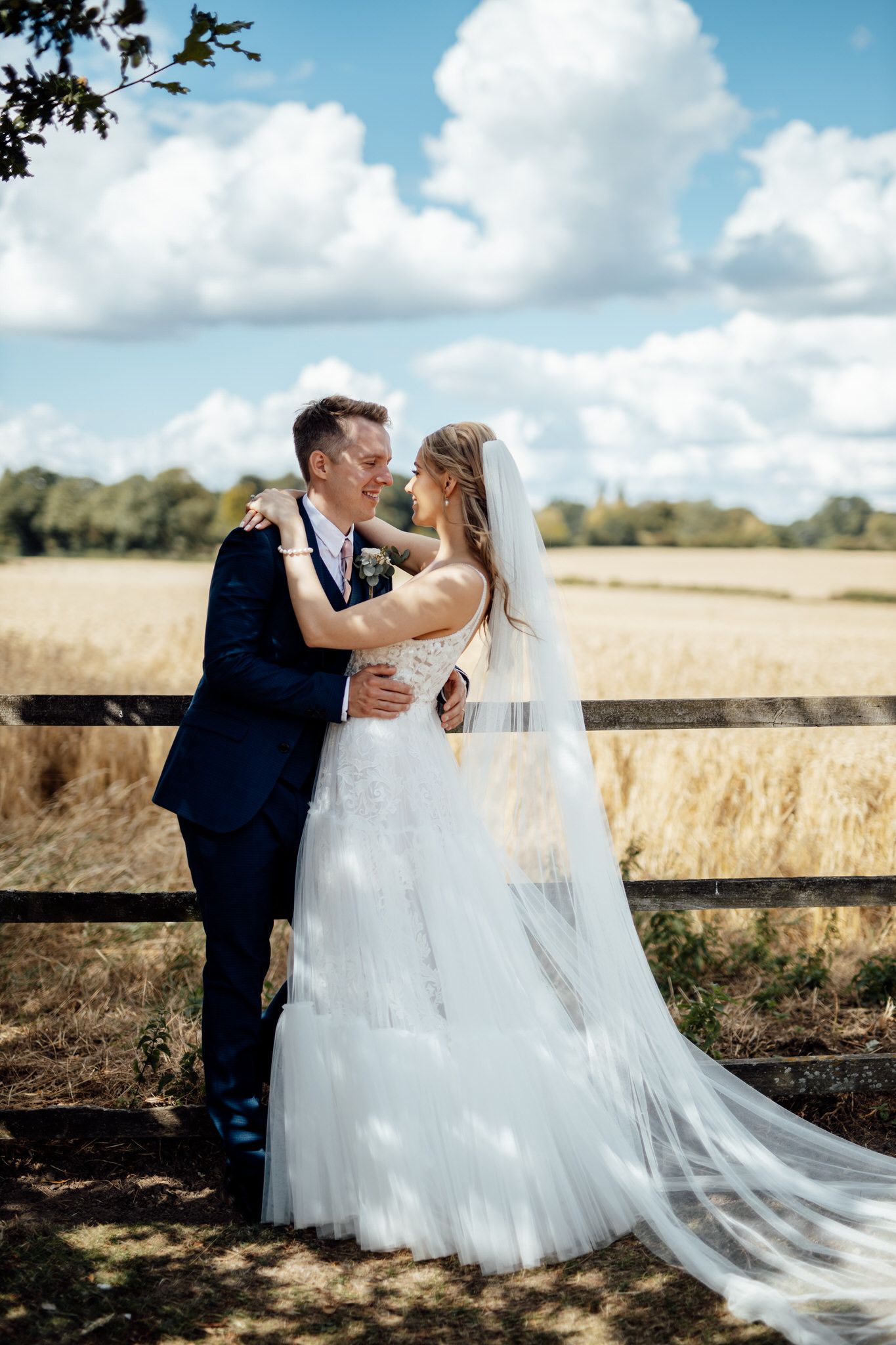 Wedding couple poses by rustic wooden fence with flowing white dress and blue sky backdrop.