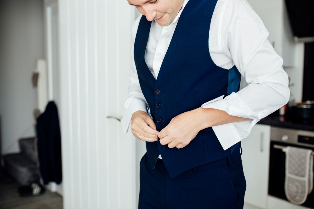 Men in navy suits and white shirts prepare formal attire indoors.