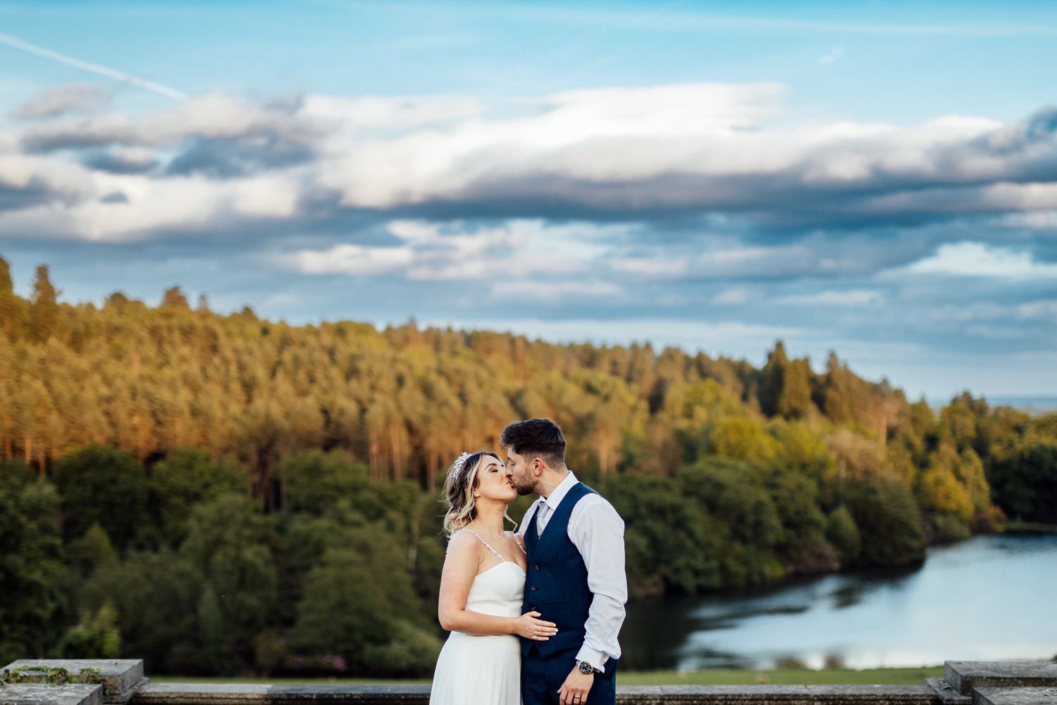 Scenic lakeside wedding photos with dramatic clouds and autumn foliage in the background.