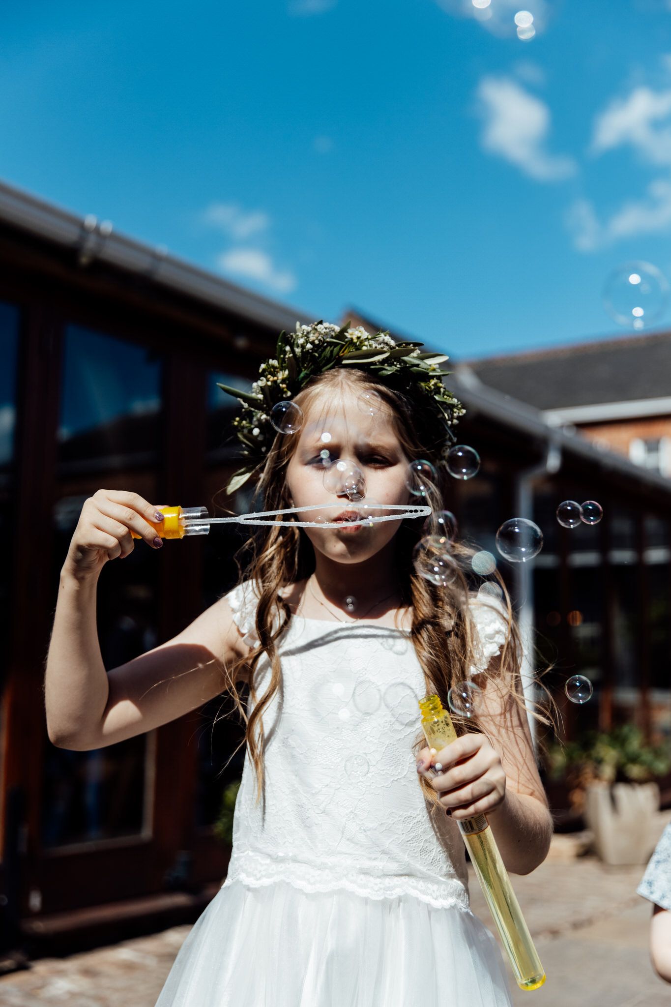 A young girl in a white dress blows bubbles outdoors on a sunny day.