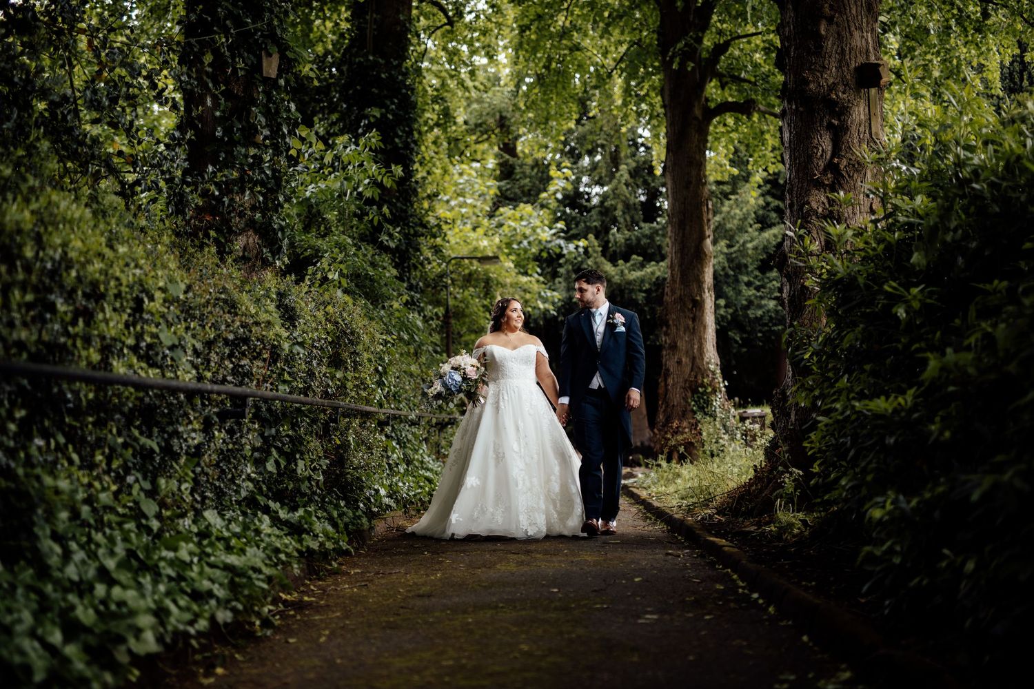 Couple walking through tree-lined pathway in formal wedding attire.