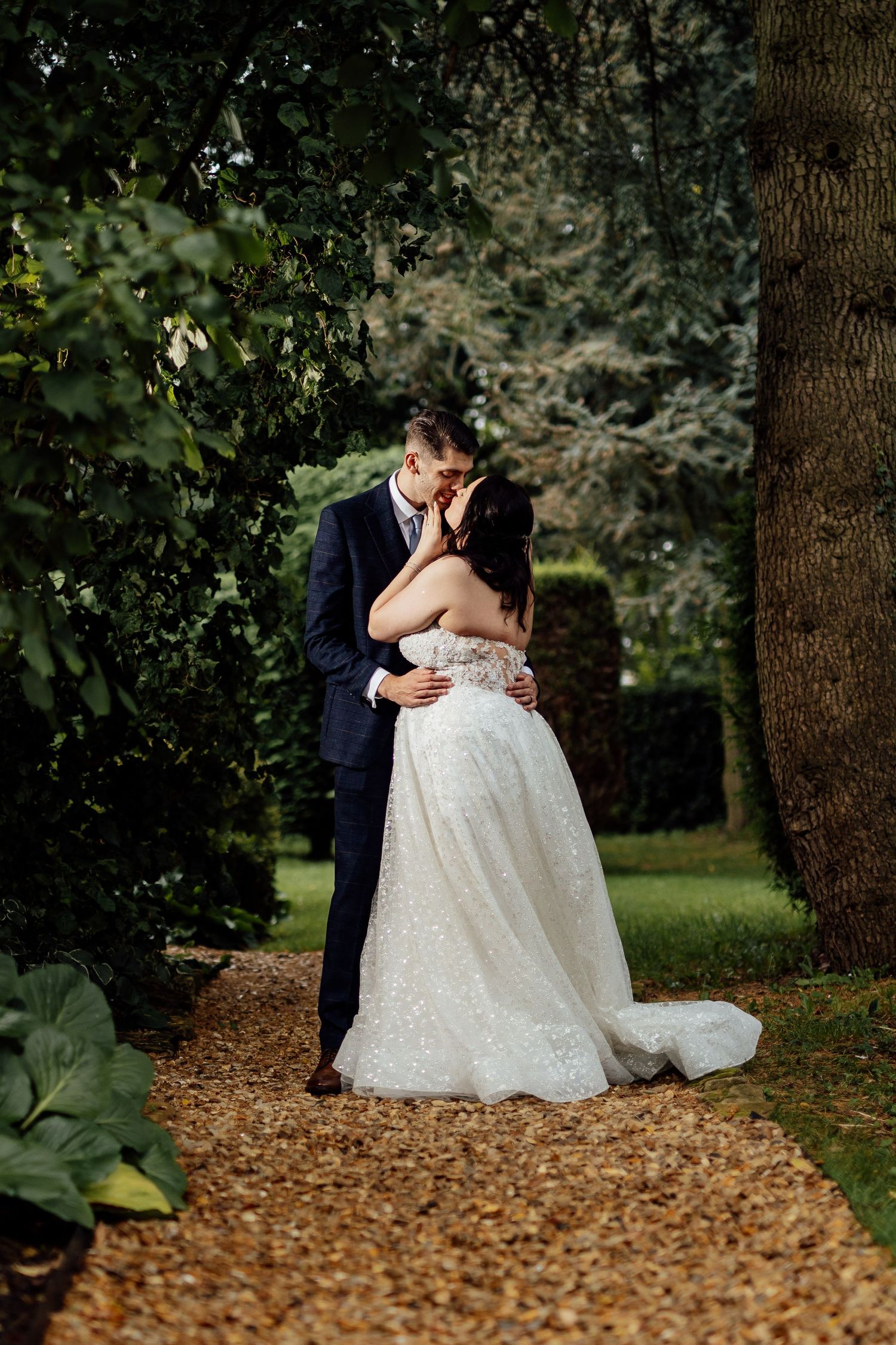 Romantic moment between newlyweds embracing on garden path surrounded by trees.