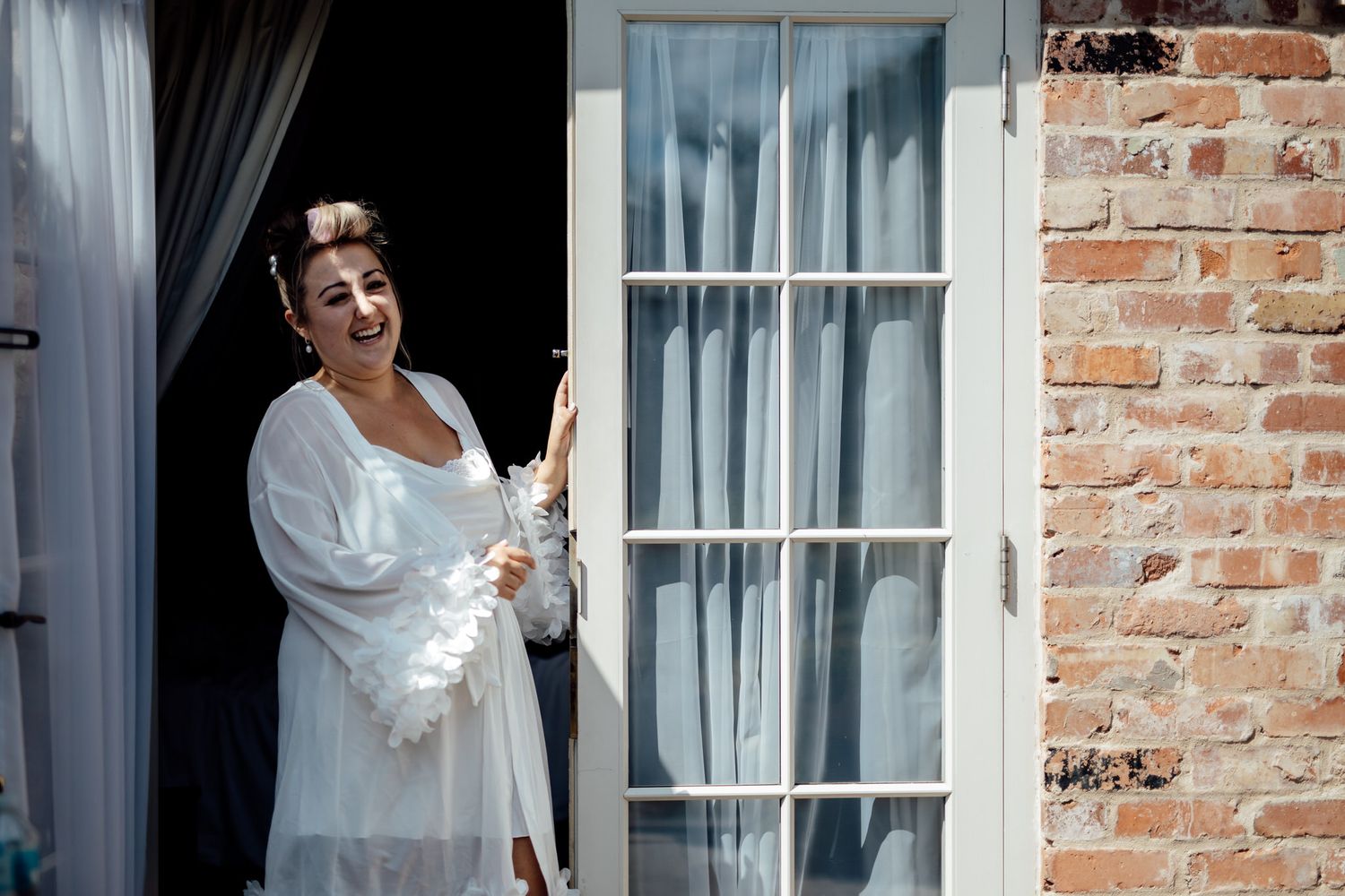 Getting ready shots through windows and doorways showing bride in white robe at rustic brick venue.