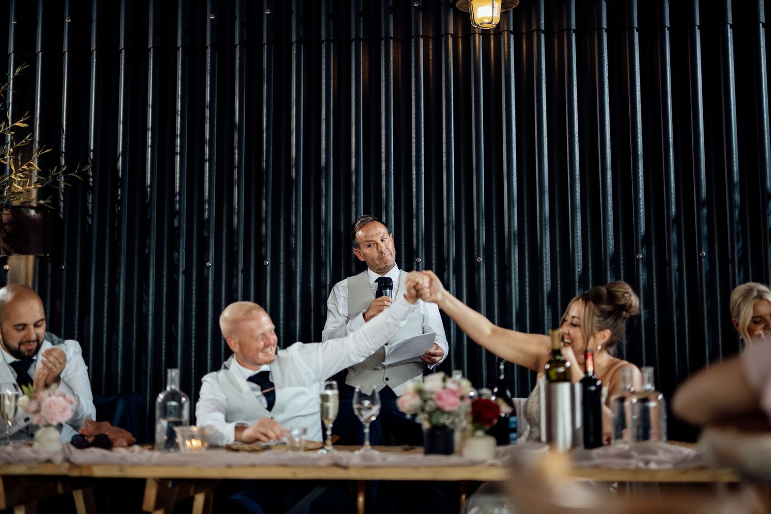Wedding reception guests seated at long tables with dark industrial-style backdrop.
