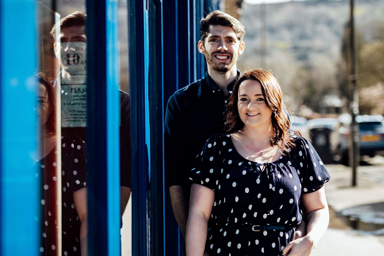 A couple poses against a bright blue wall on a sunny day in casual outfits.