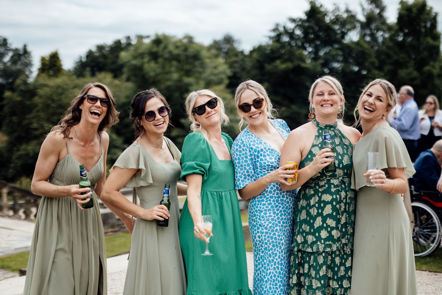 Bridesmaids in sage green and floral dresses laugh together while holding drinks at outdoor reception.
