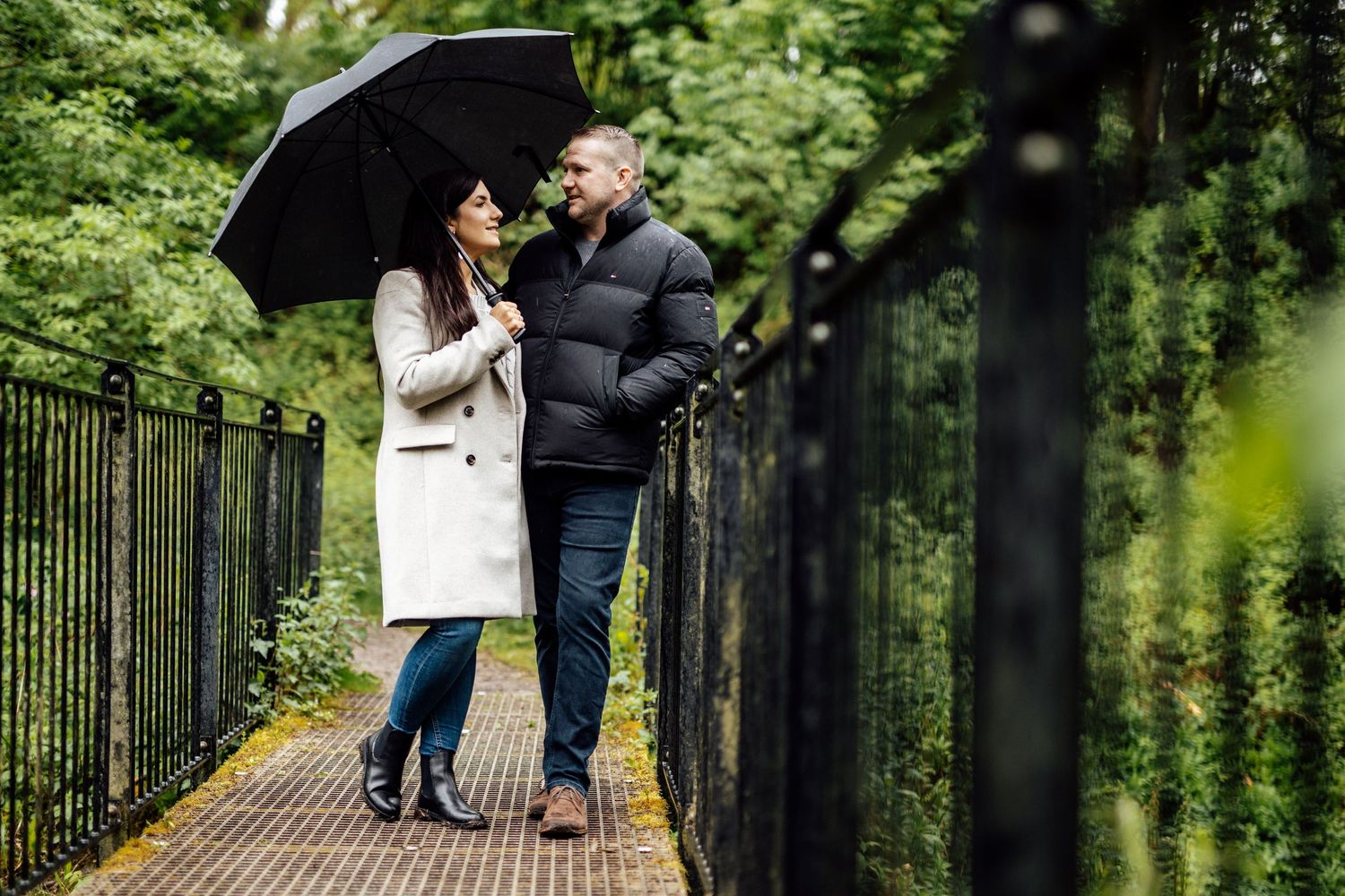 A couple walks along an iron-fenced pathway through a lush green park.