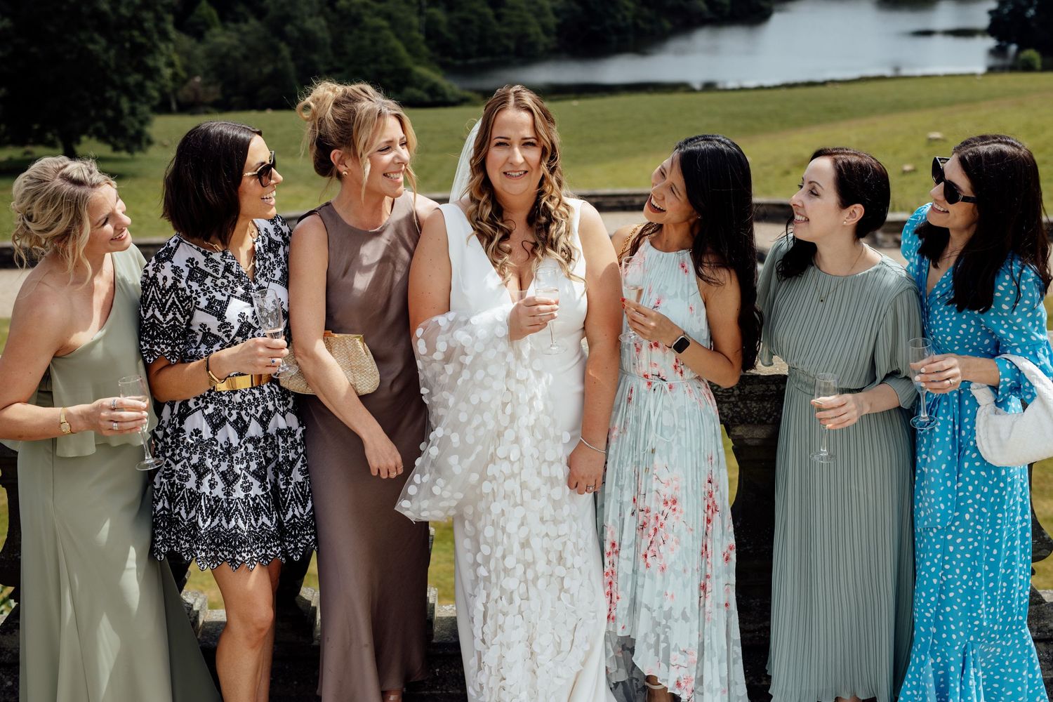 Wedding party in pastel and floral dresses posing together by scenic lake view.