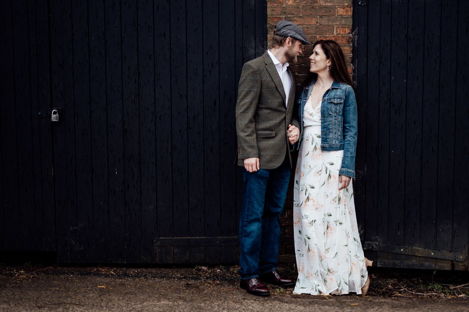 A couple stands against a dark wooden wall in casual wedding attire.
