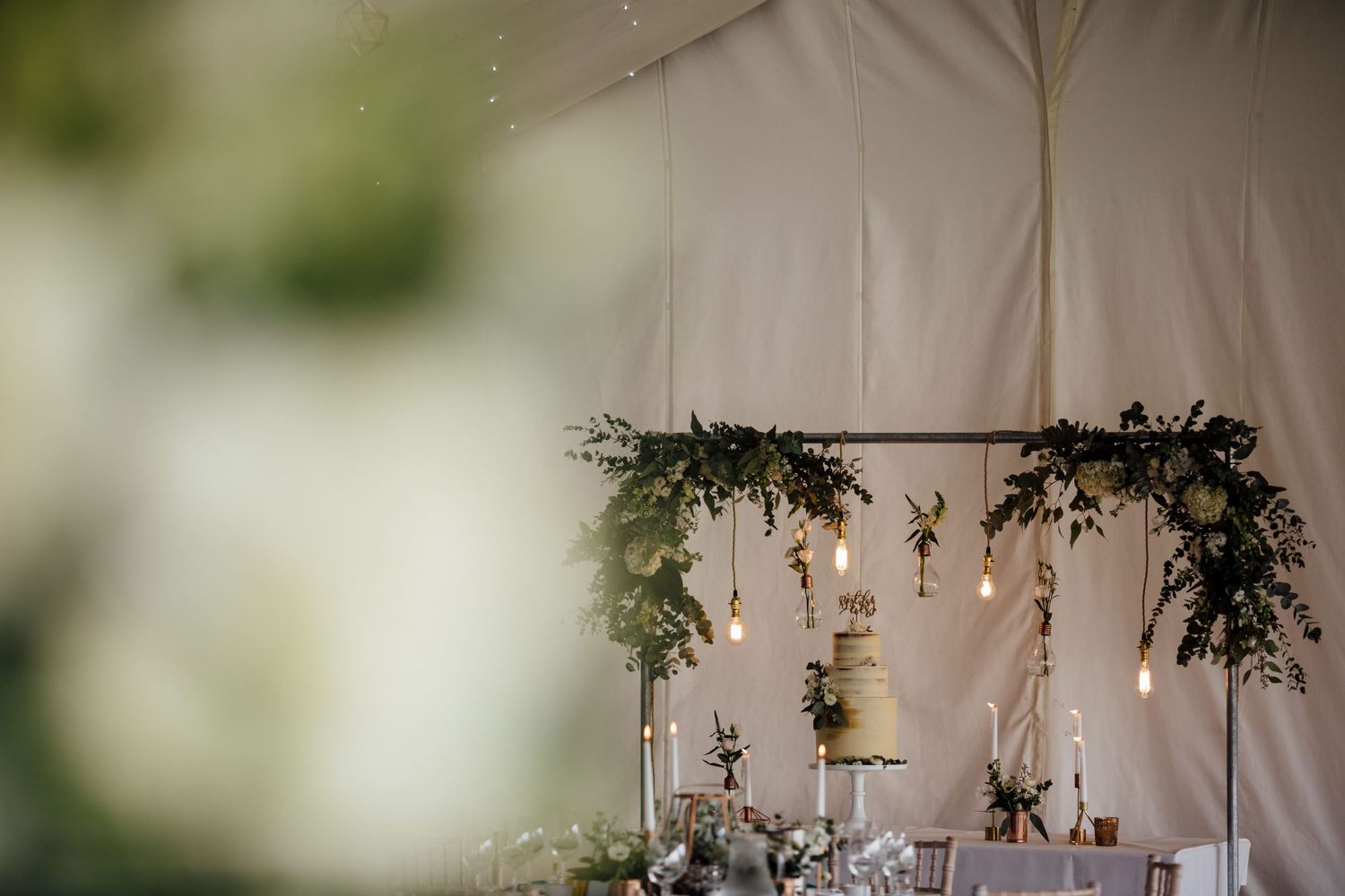 Wedding reception decor featuring hanging greenery and floral arrangements against white fabric.