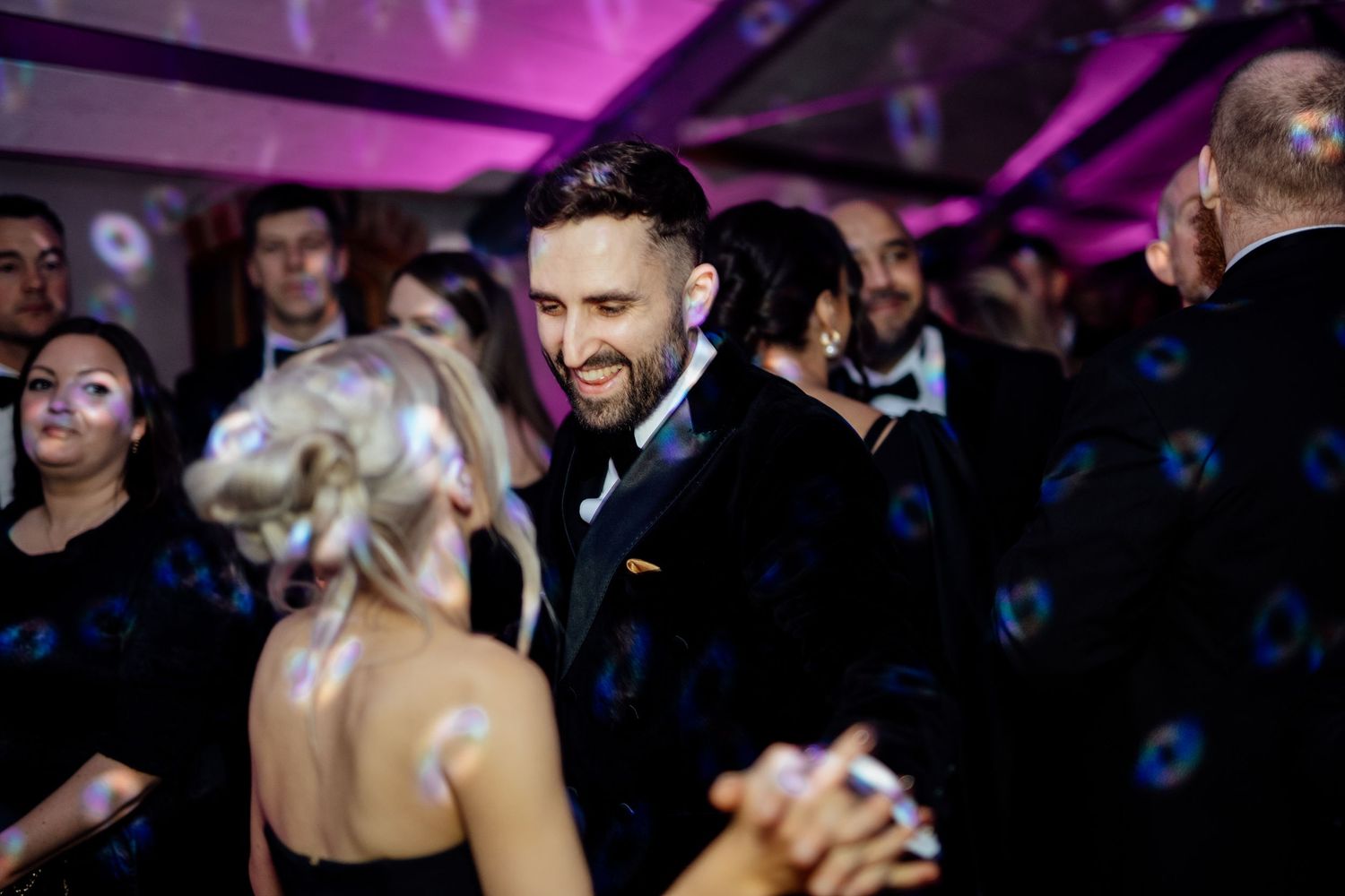 Wedding guests dance and celebrate on a dimly lit dance floor with purple lighting and festive atmosphere.