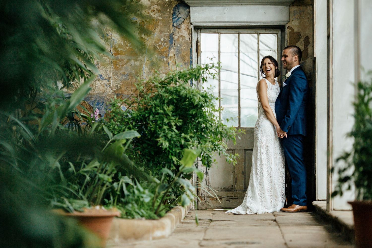 A couple in wedding attire stands in a romantic stone archway surrounded by lush greenery and natural light.