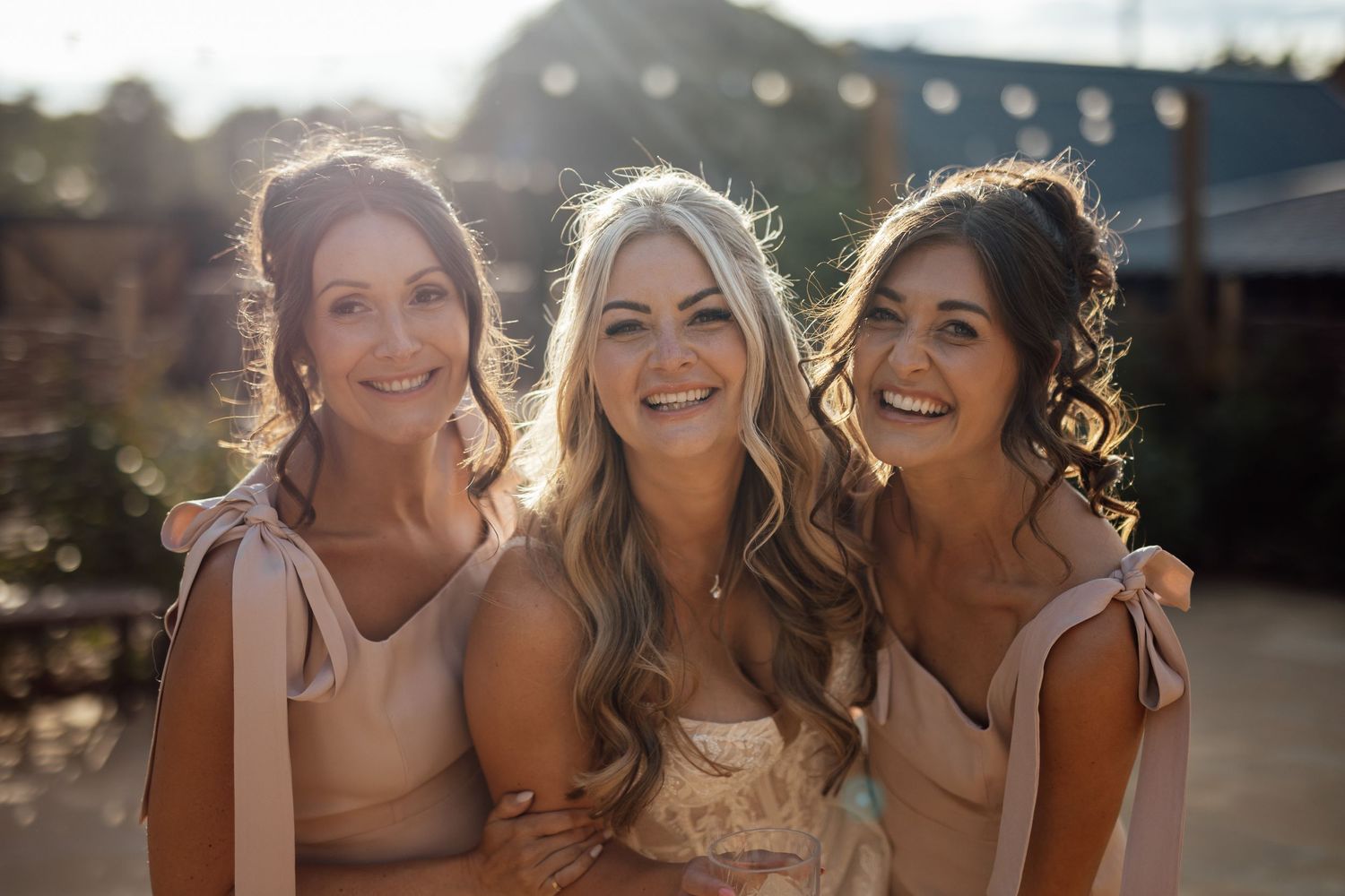 Smiling bride poses with two bridesmaids in blush dresses at sunny outdoor wedding venue.