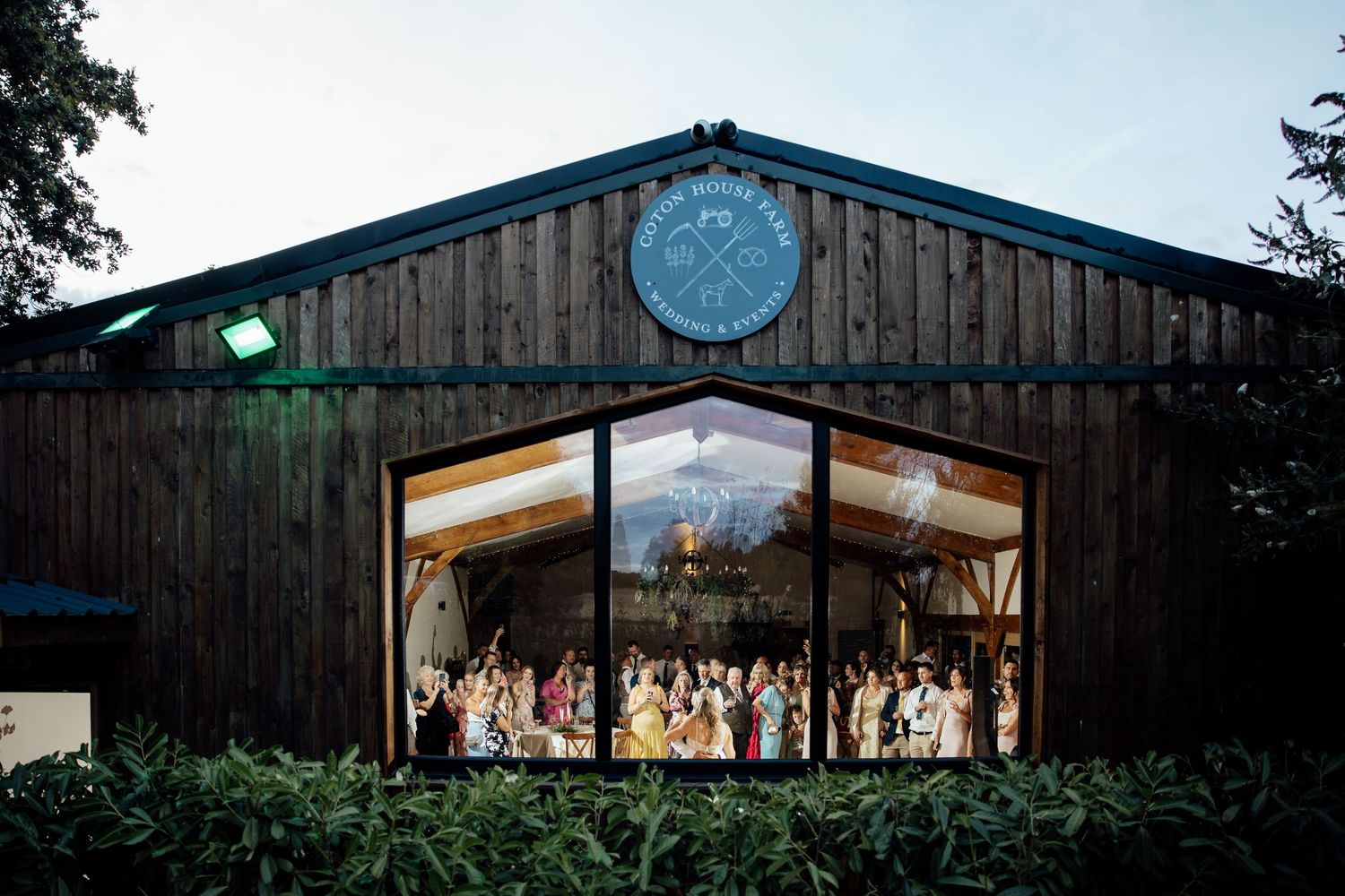 Wedding guests gathered inside warmly lit rustic barn venue photographed through large glass windows at dusk.