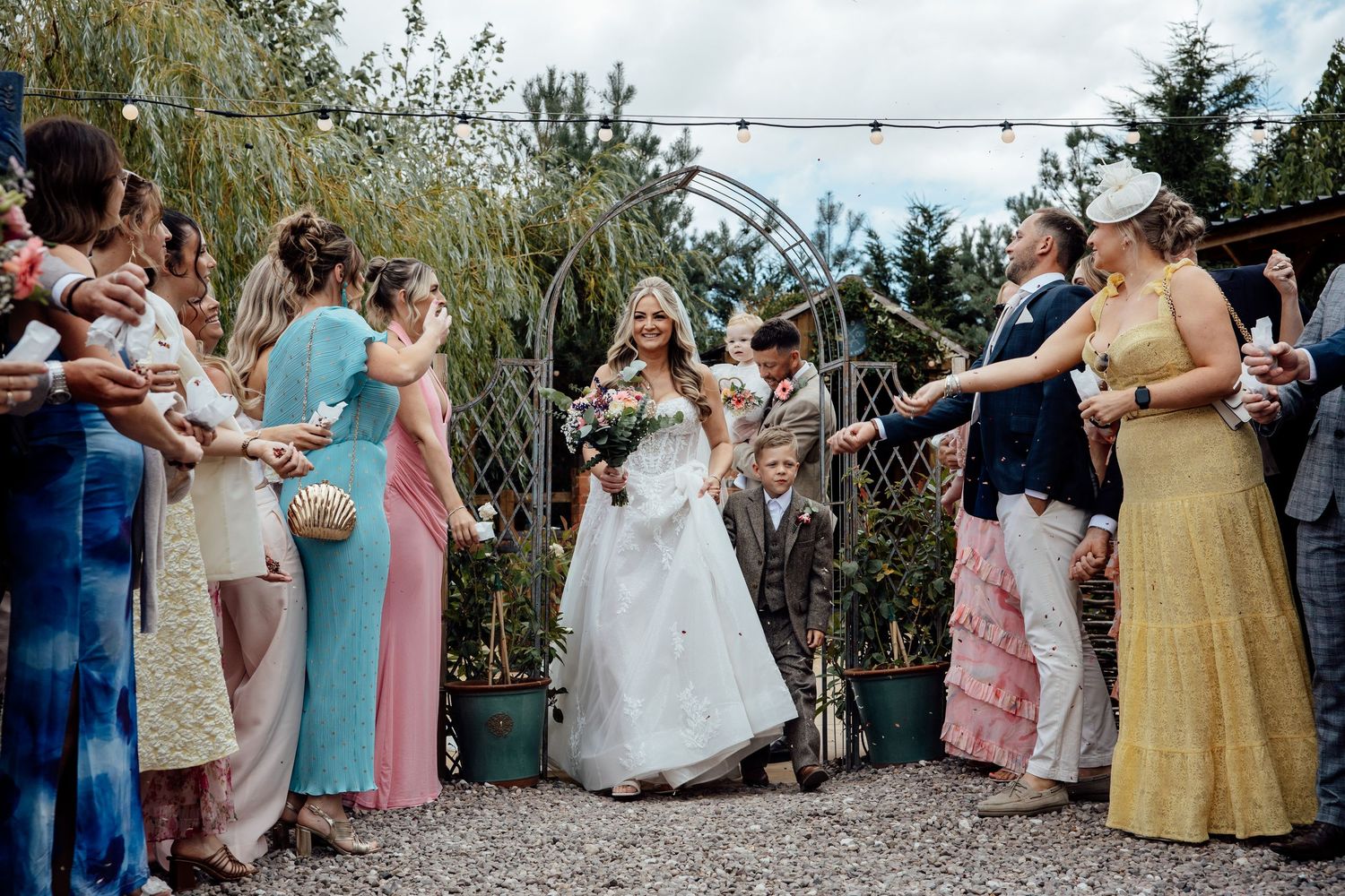 A radiant bride walks through a confetti tunnel of cheering wedding guests on a gravel path.