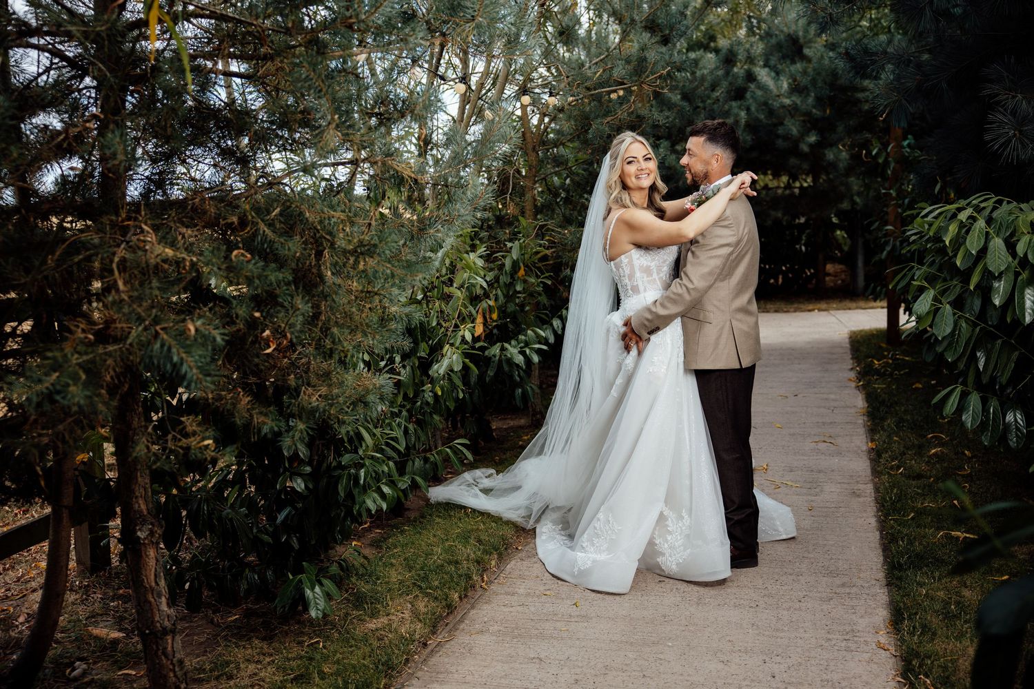 Bride and groom embracing on garden path surrounded by lush green trees.
