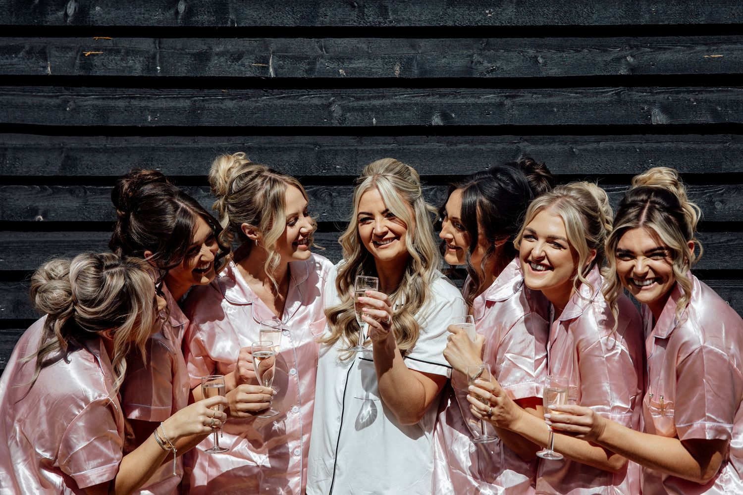 Bride in white robe laughing with bridesmaids in pink satin robes holding champagne glasses during wedding morning prep.