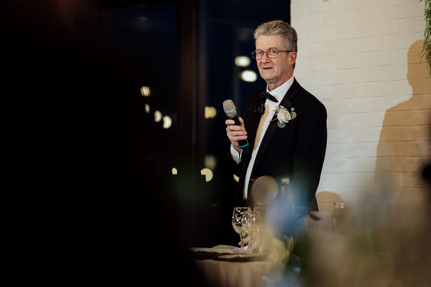 Elegant groom in black tuxedo delivers a heartfelt wedding speech holding a microphone at the reception.