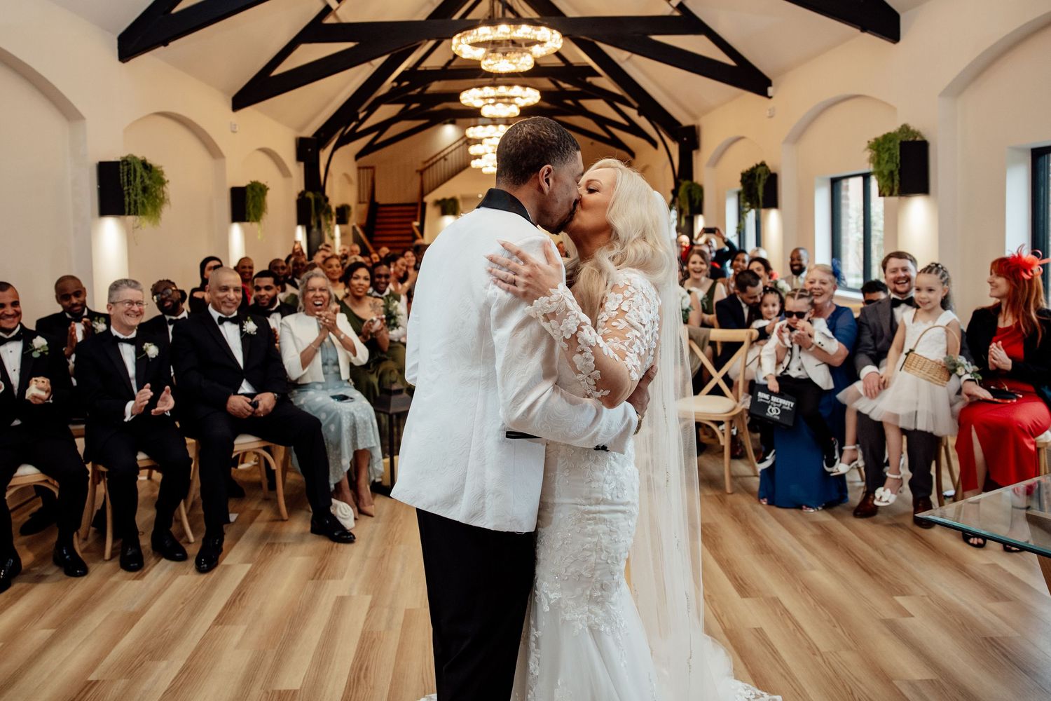 Bride and groom share first kiss at wedding altar as guests cheer in elegant venue.