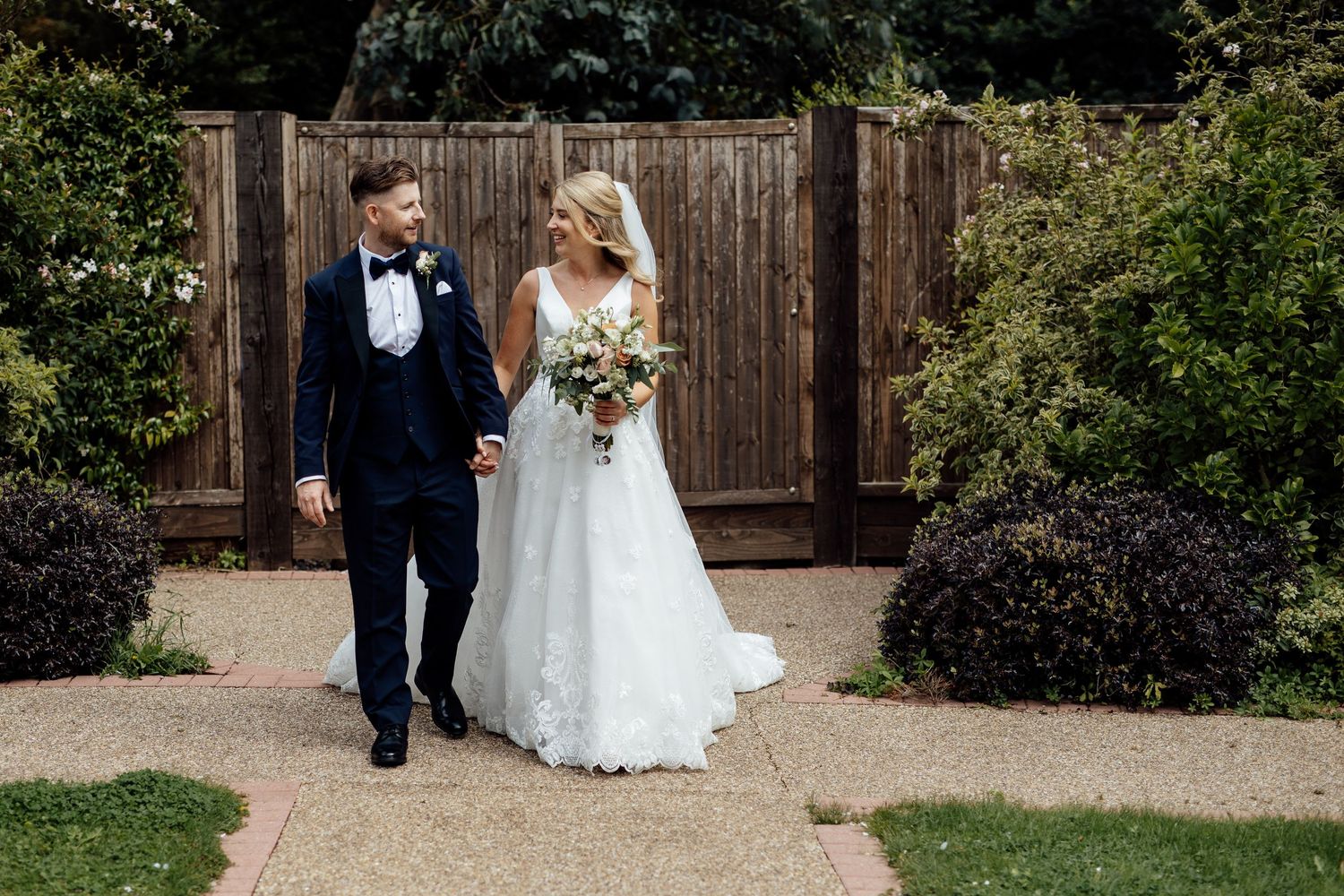 Wedding couple walking through garden path with wooden fence backdrop.