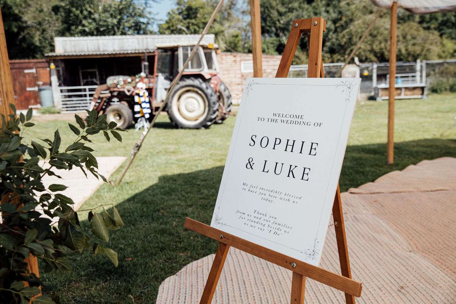 Wedding welcome sign for Sophie and Luke displayed on wooden easel with tractor in rustic farm background.