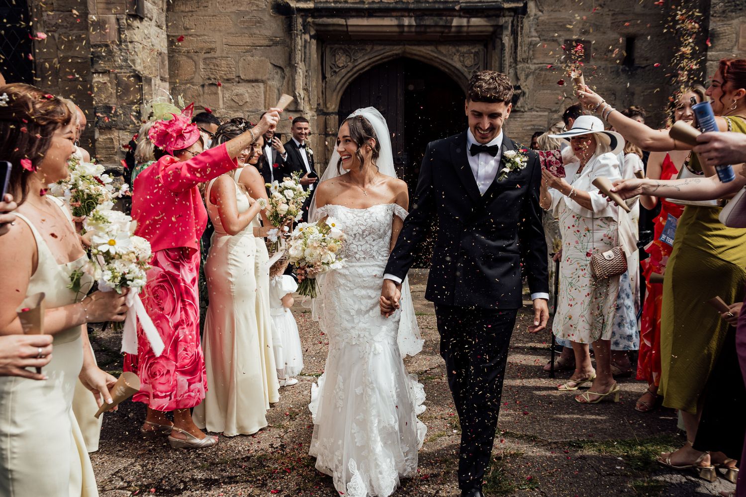 Wedding celebration with guests throwing confetti in front of stone church walls.