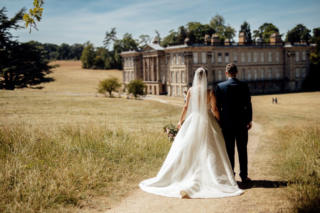 A couple in wedding attire walks through a scenic meadow with historic buildings in the background.