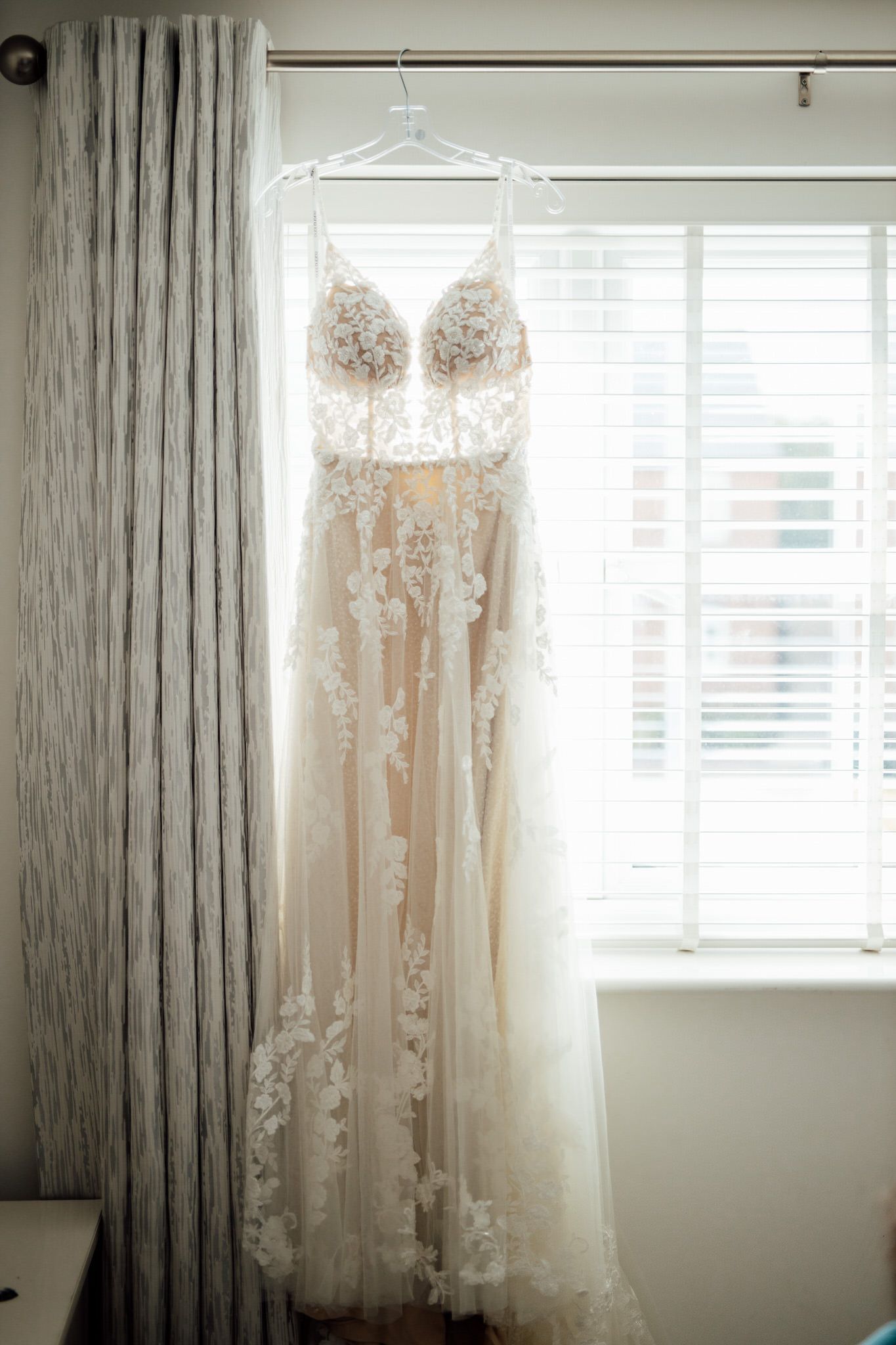 Delicate lace wedding dress hangs by window with soft natural lighting.