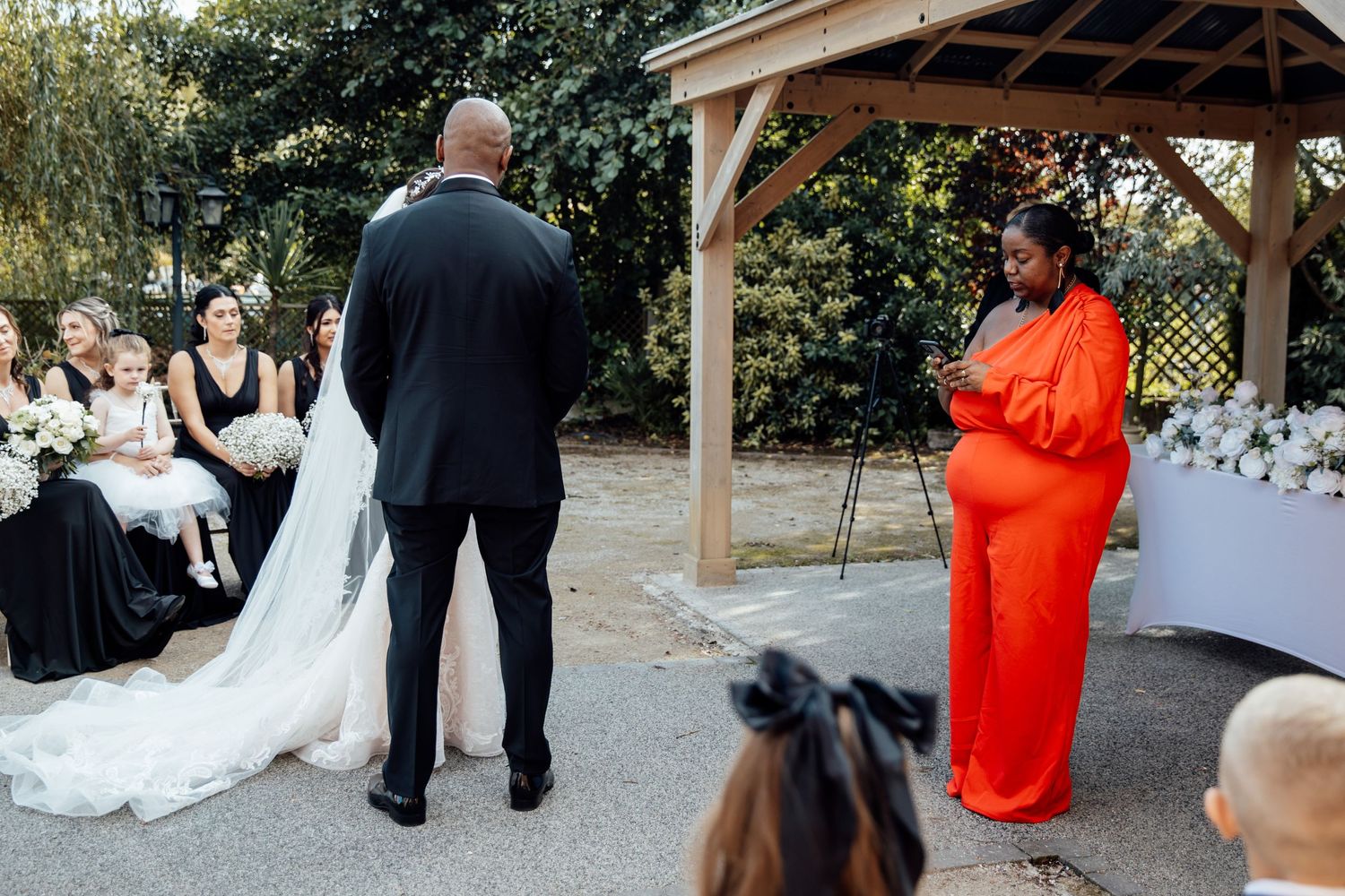 Outdoor wedding ceremony under wooden gazebo with guests gathered around.