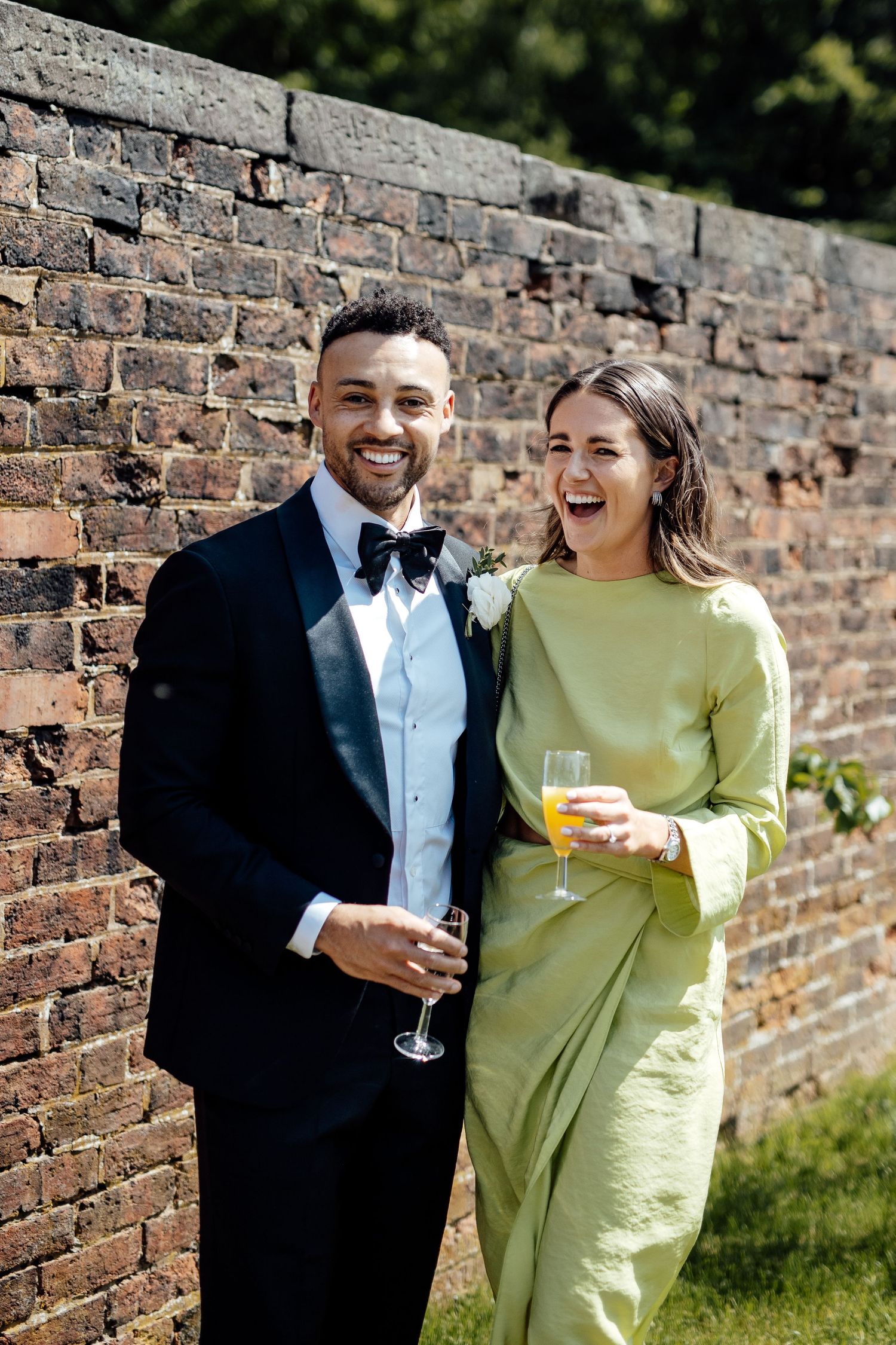 Couple in formal attire shares laughs against brick wall during wedding celebration.