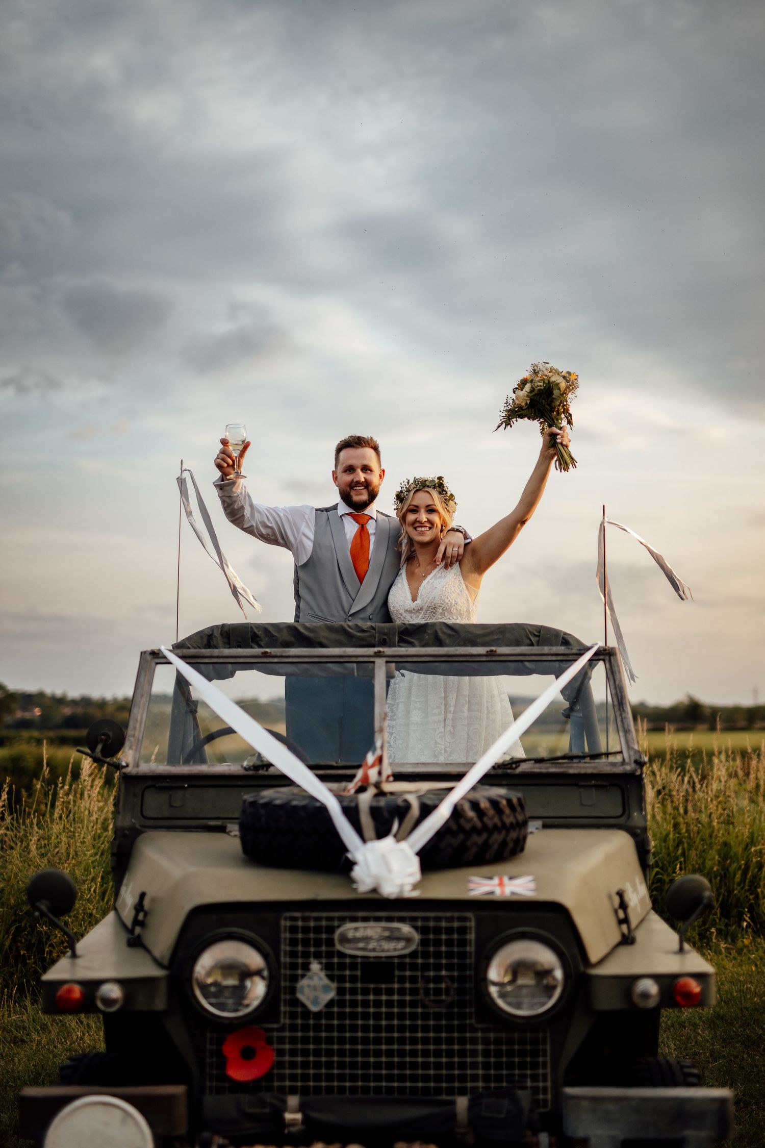 Newlyweds celebrate atop vintage Land Rover decorated with white ribbons at sunset.