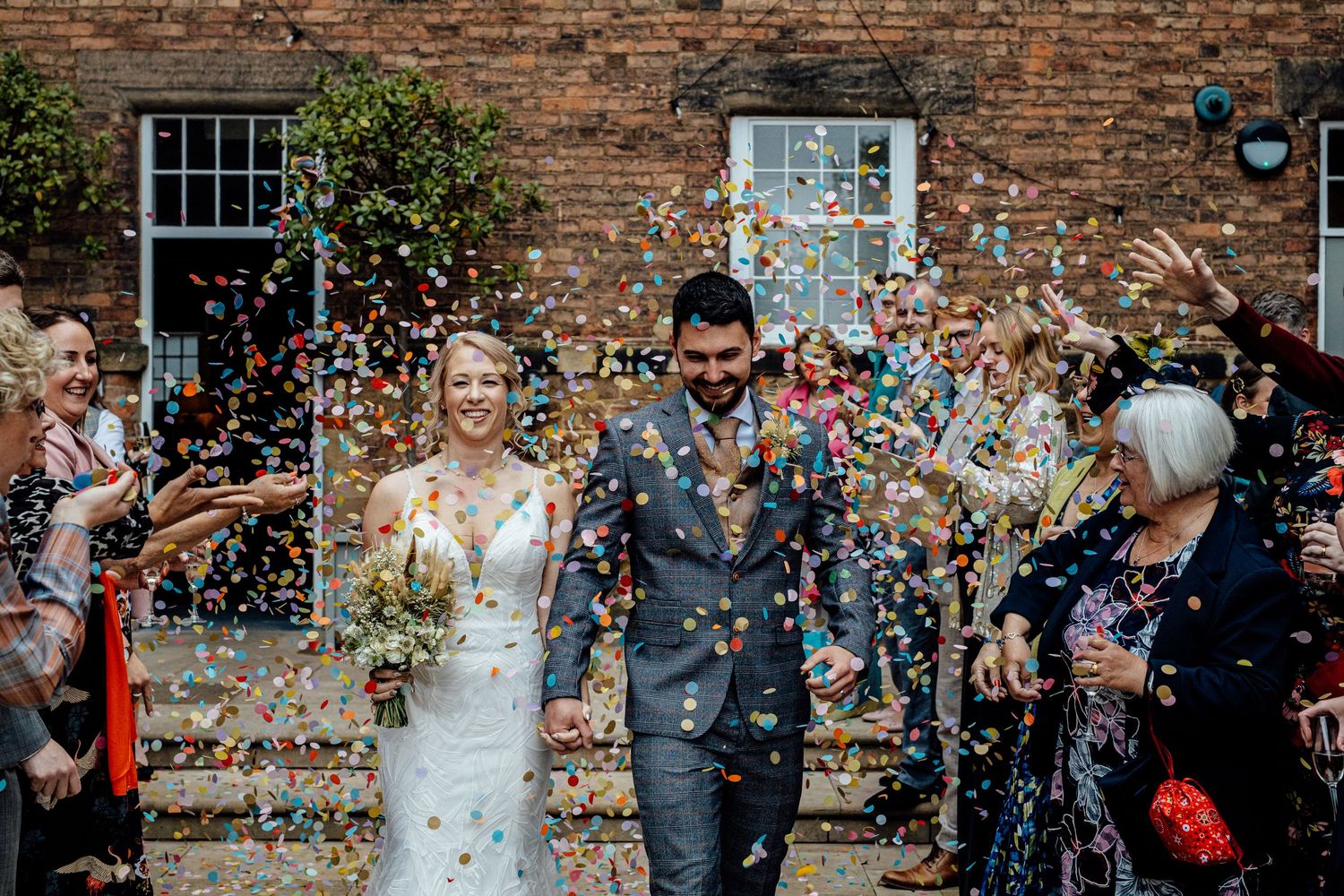 Wedding celebration with confetti being thrown against a historic brick building backdrop.