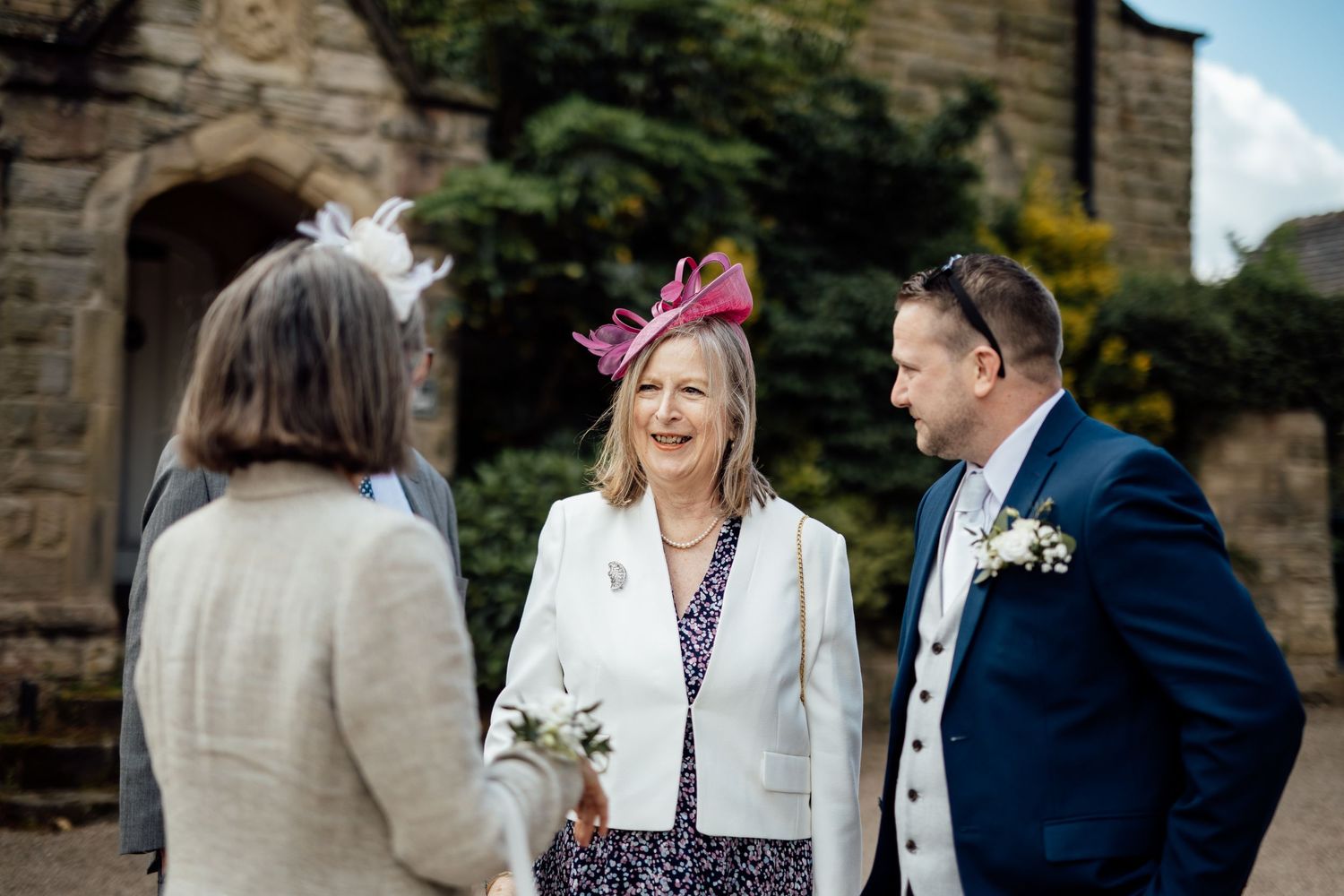 Wedding guests mingling outdoors near stone building on sunny day.