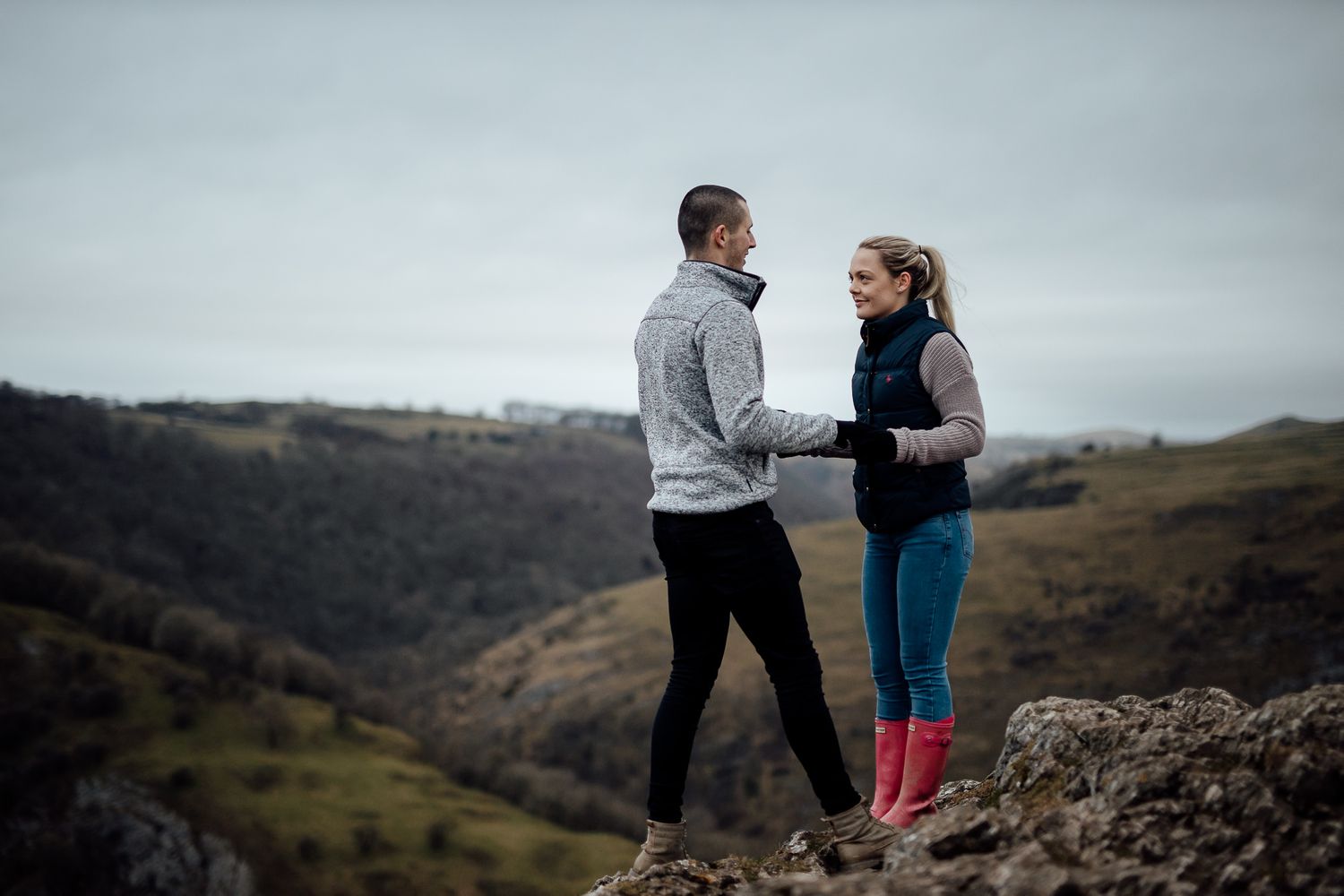 A couple stands together against a moody mountain landscape backdrop during an engagement photoshoot.