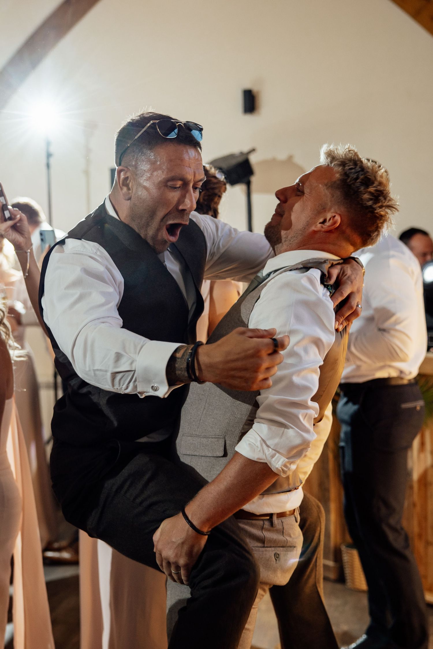 Two male wedding guests laugh and playfully wrestle on the dance floor at a reception.